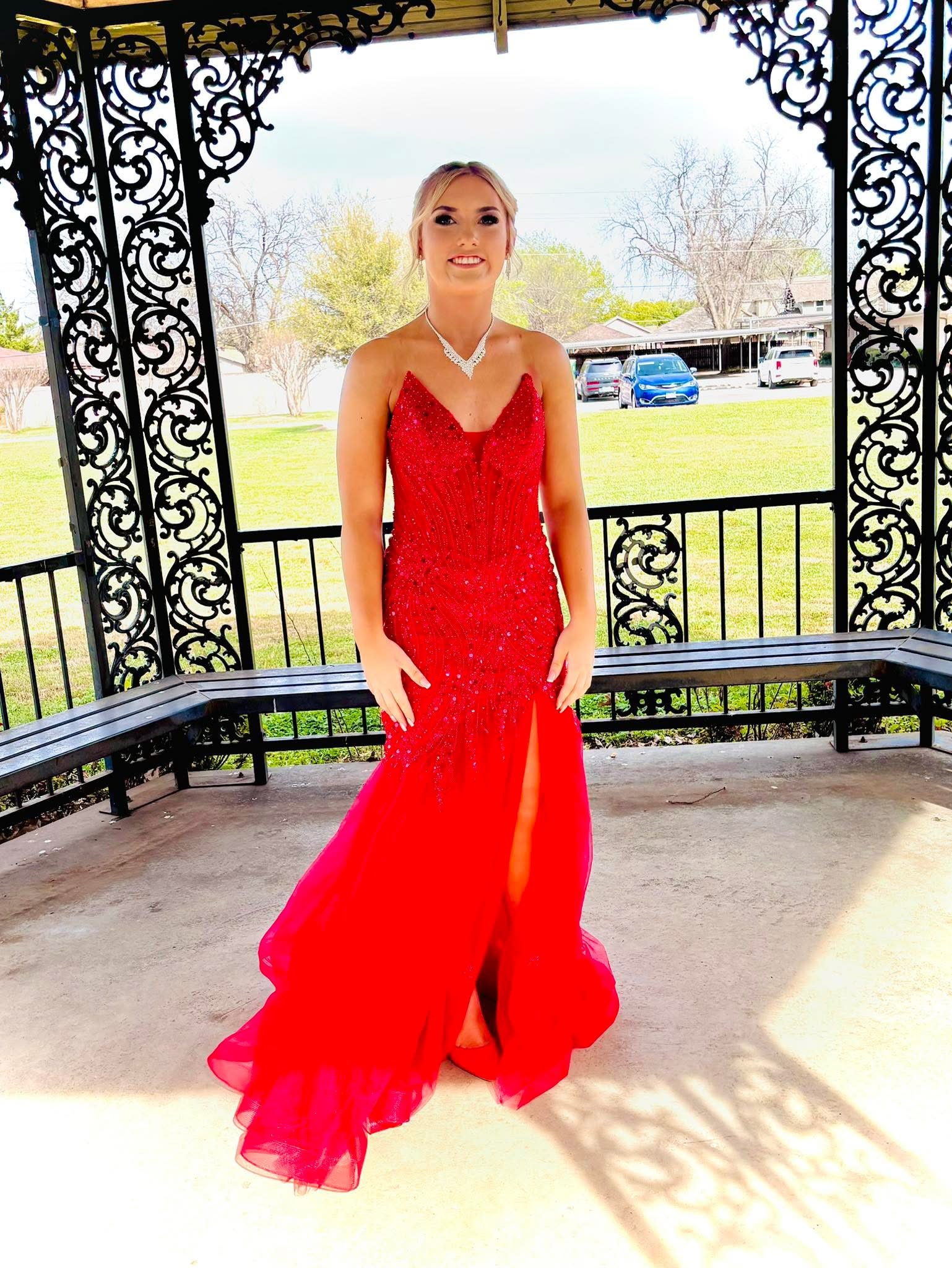 A young woman posing in a floor-length red sequined formal gown inside an ornate black wrought-iron gazebo, with a park setting in the background.