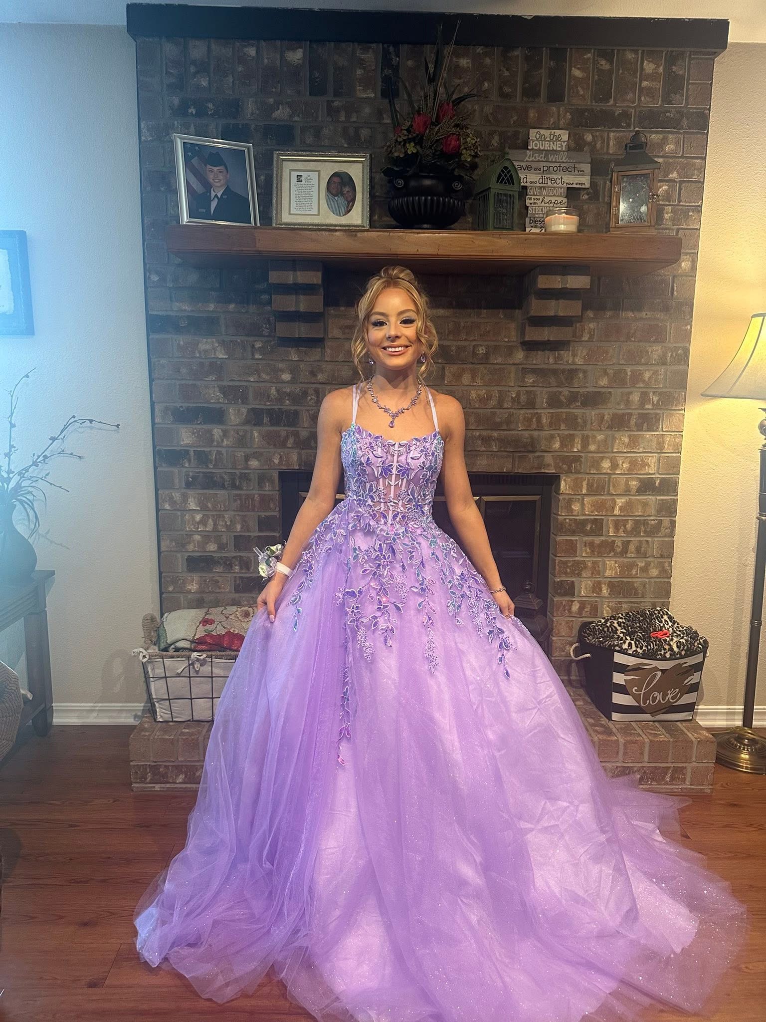 A smiling young woman in a lavender formal gown with a lace bodice and tulle skirt posing in front of a brick fireplace.