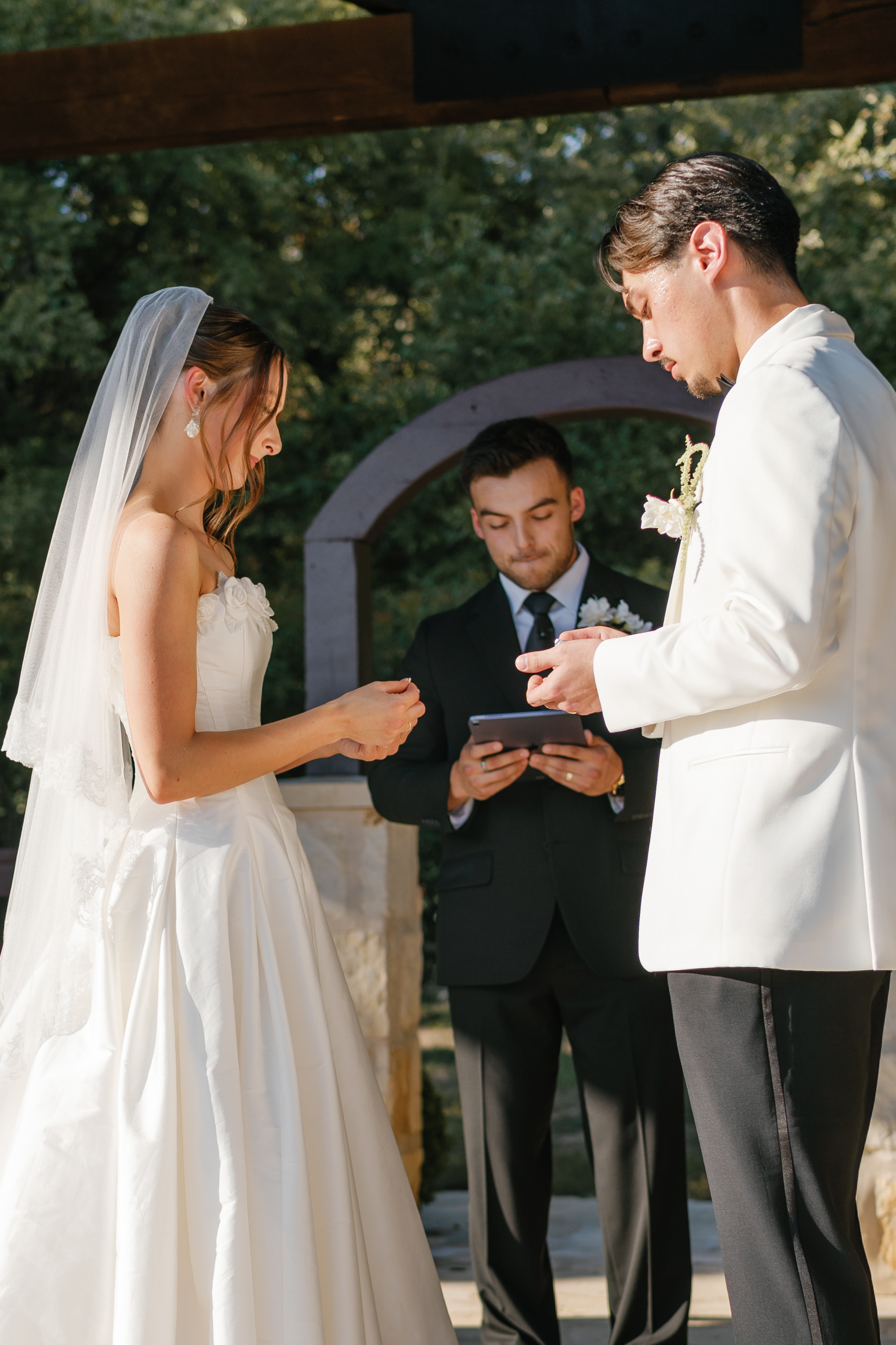 A bride and groom exchanging vows during an outdoor wedding ceremony with an officiant holding a tablet between them.