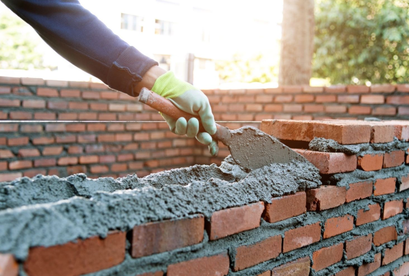Bricklaying with trowel applying mortar to red brick wall
