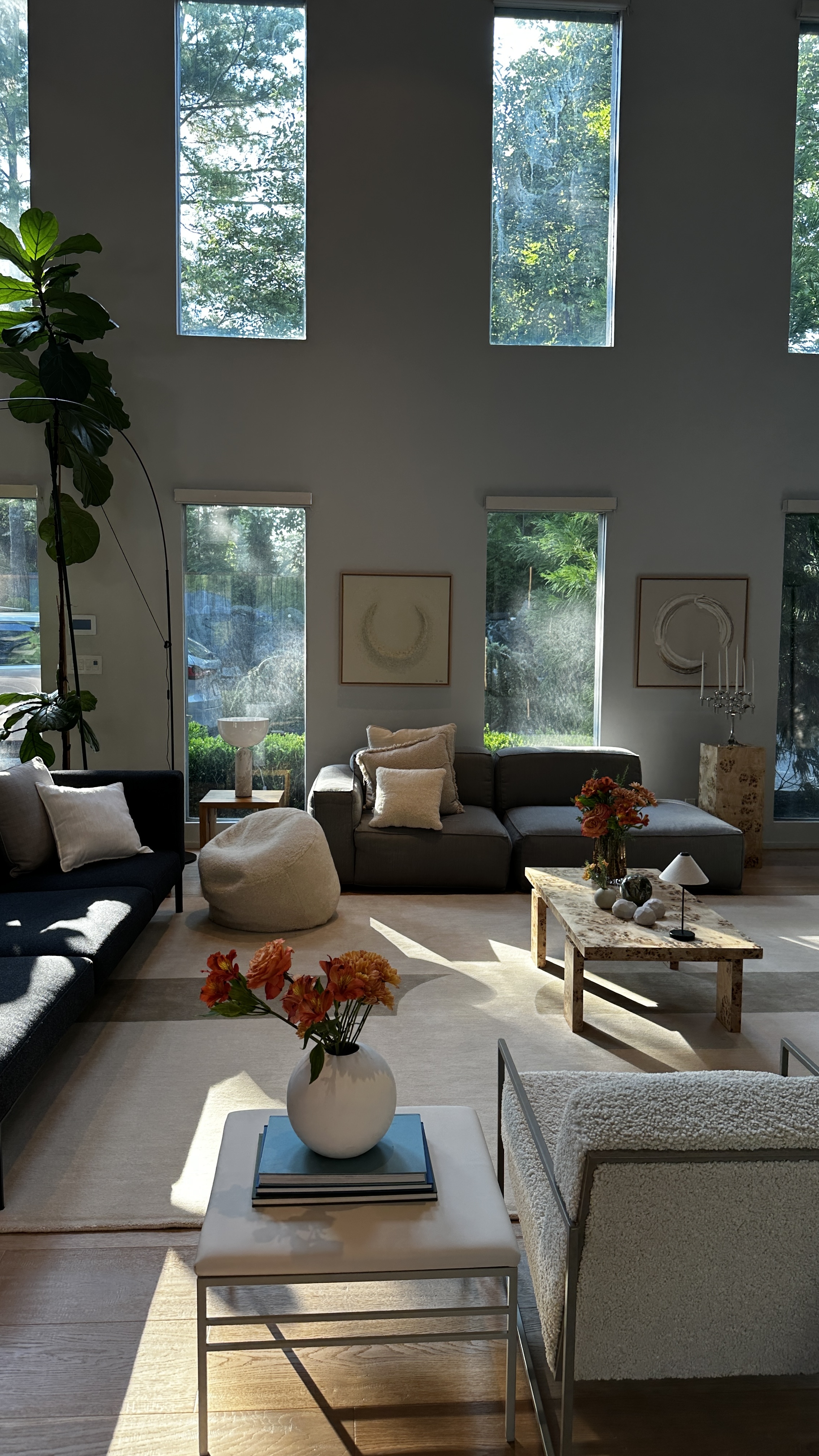 Double-height living room with tall slot clerestory windows onto trees, a charcoal modular sectional, bouclé beanbag, burl stone coffee table with orange blooms and framed circular relief artworks