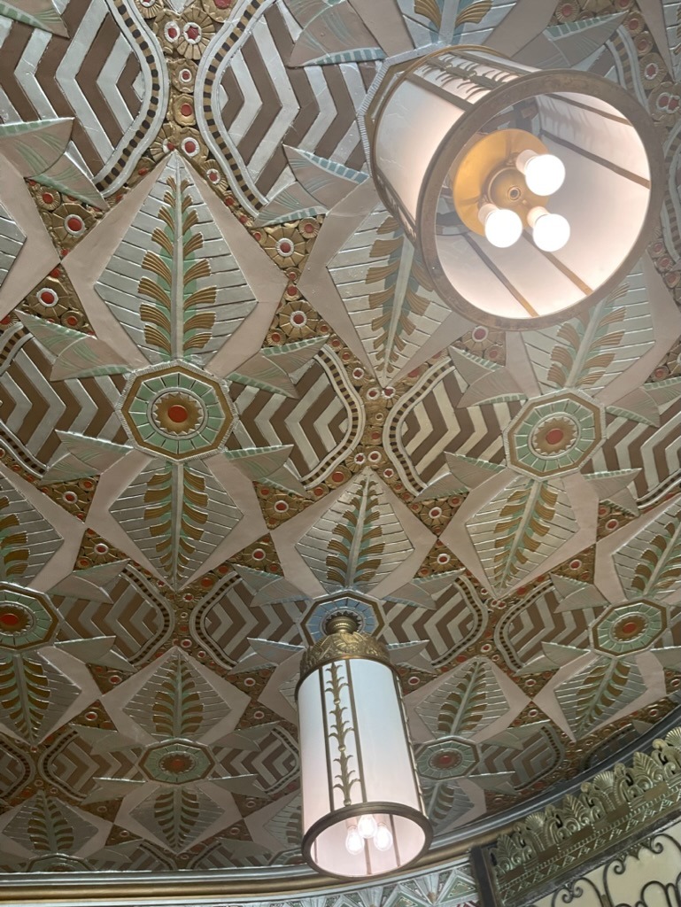Low-angle view of the ornate Art Deco lobby ceiling with a repeating diamond and octagonal medallion pattern in gold, cream and green, and two brass cylinder pendant lights with exposed bulbs