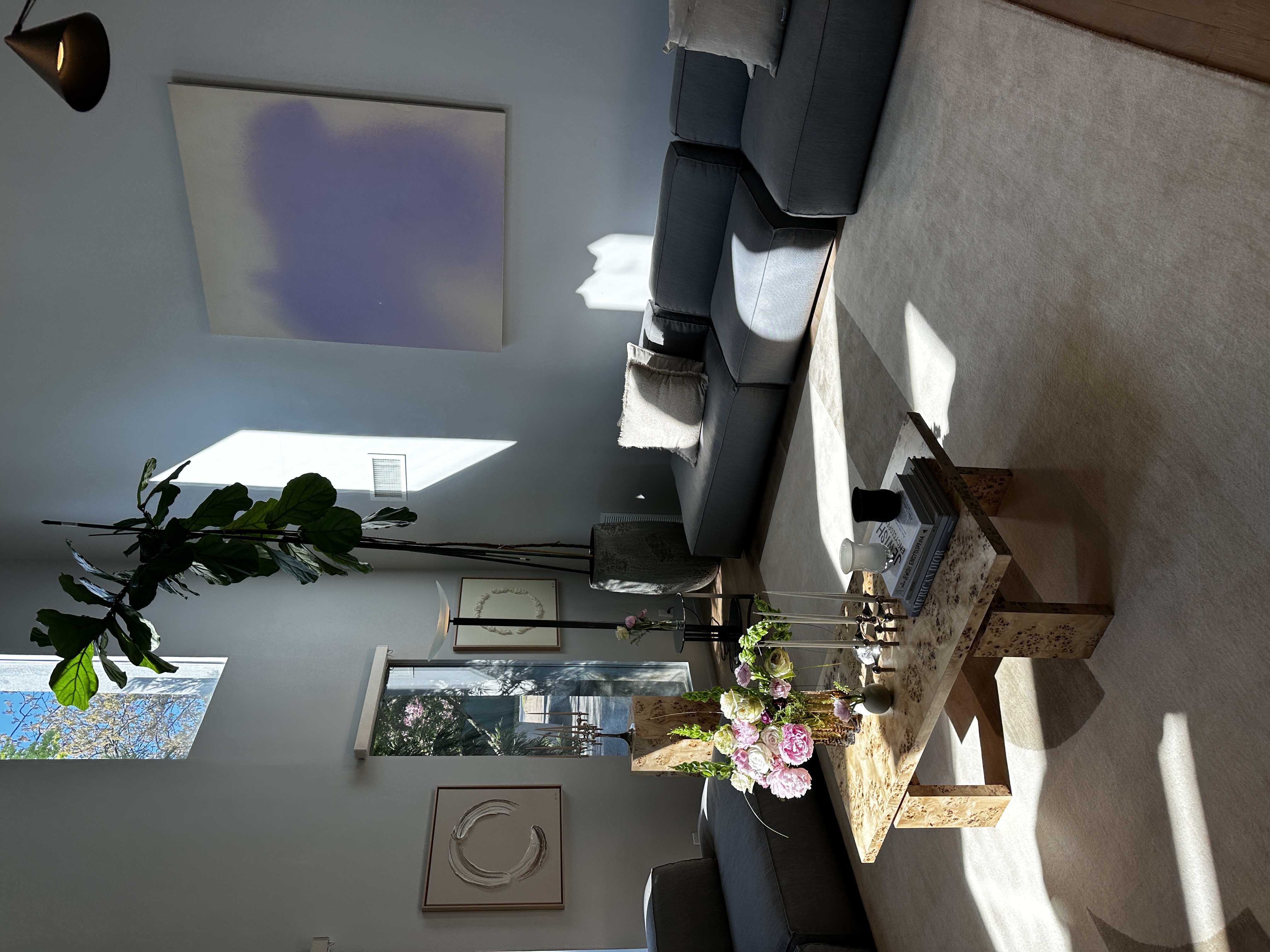 Living room corner with a large lilac abstract canvas, charcoal modular sectional, burl stone coffee table with peonies, tall fiddle-leaf fig and slot windows onto blossom