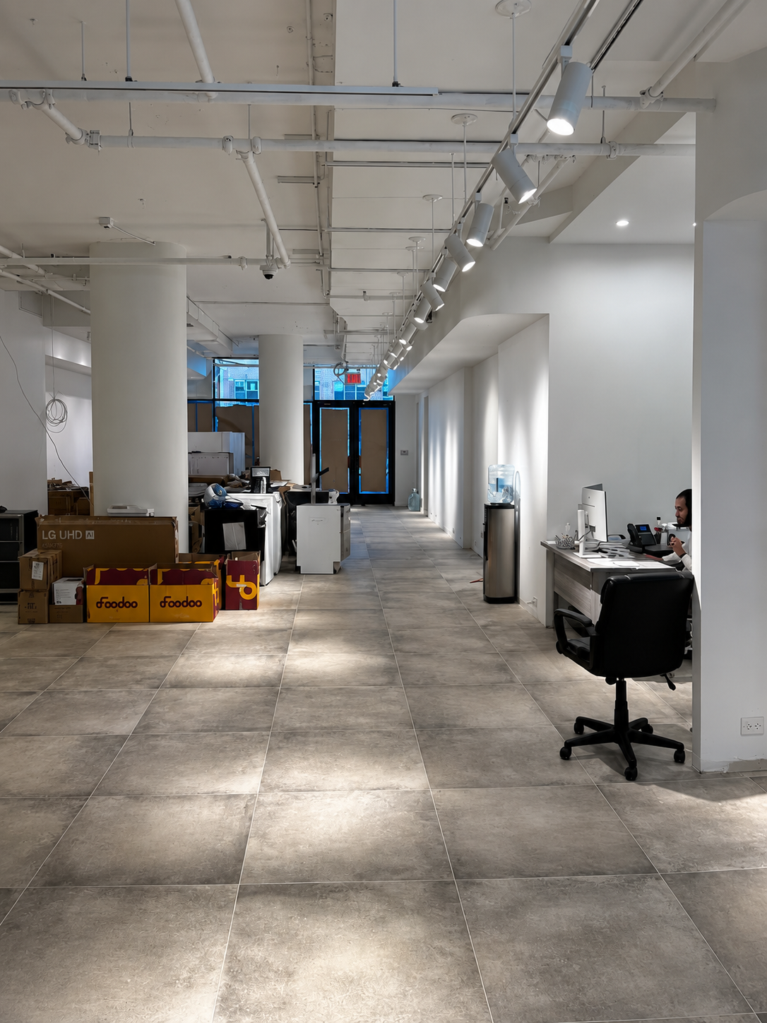 Showroom during fit-out with track spotlights on exposed services, appliance cartons and crates along one side and a staff desk with computer in a lit alcove