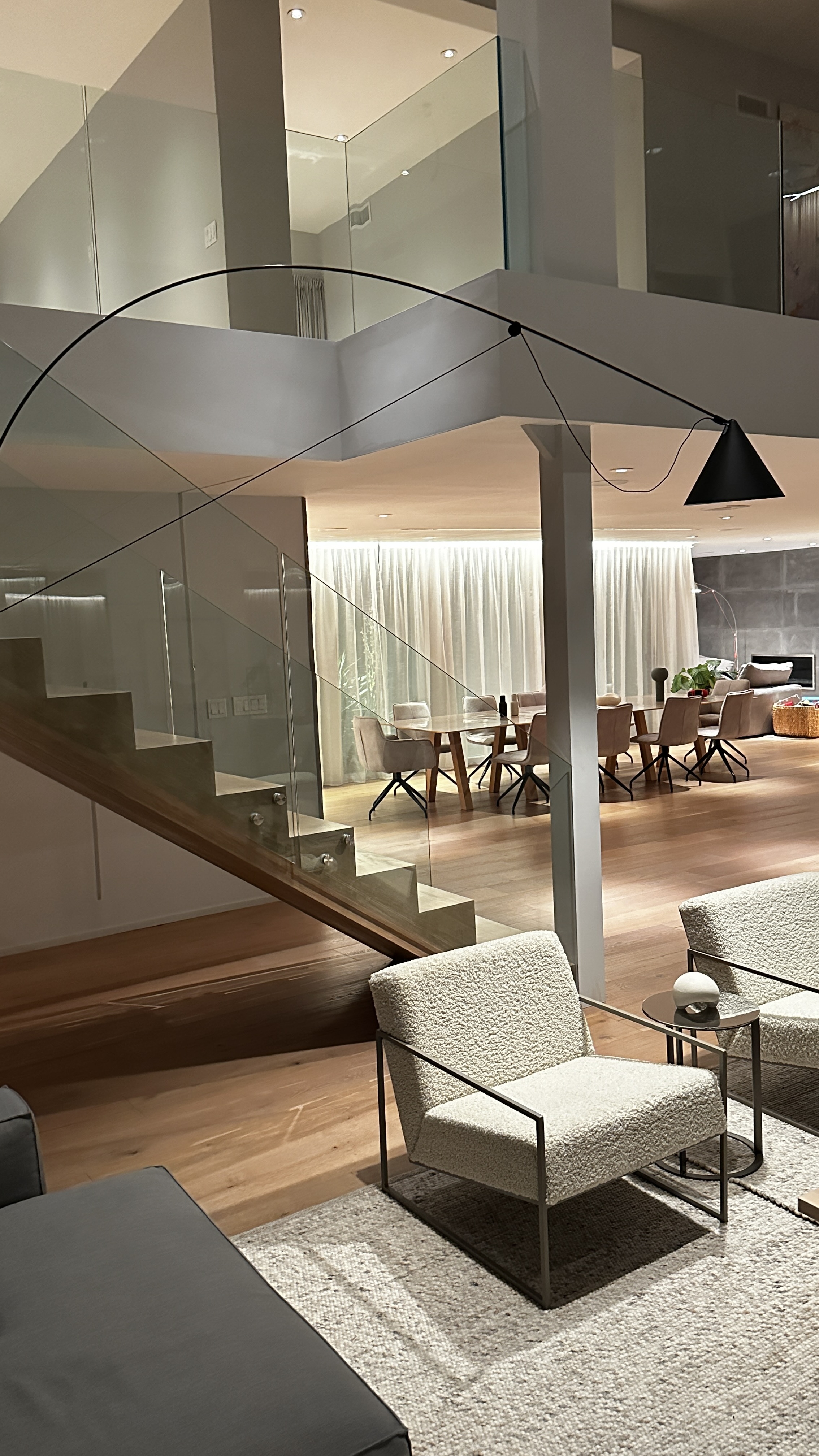 Timber and glass staircase with a frameless balustrade, a black cone pendant on a long arm, and the curtained dining room with oak table and upholstered chairs beyond