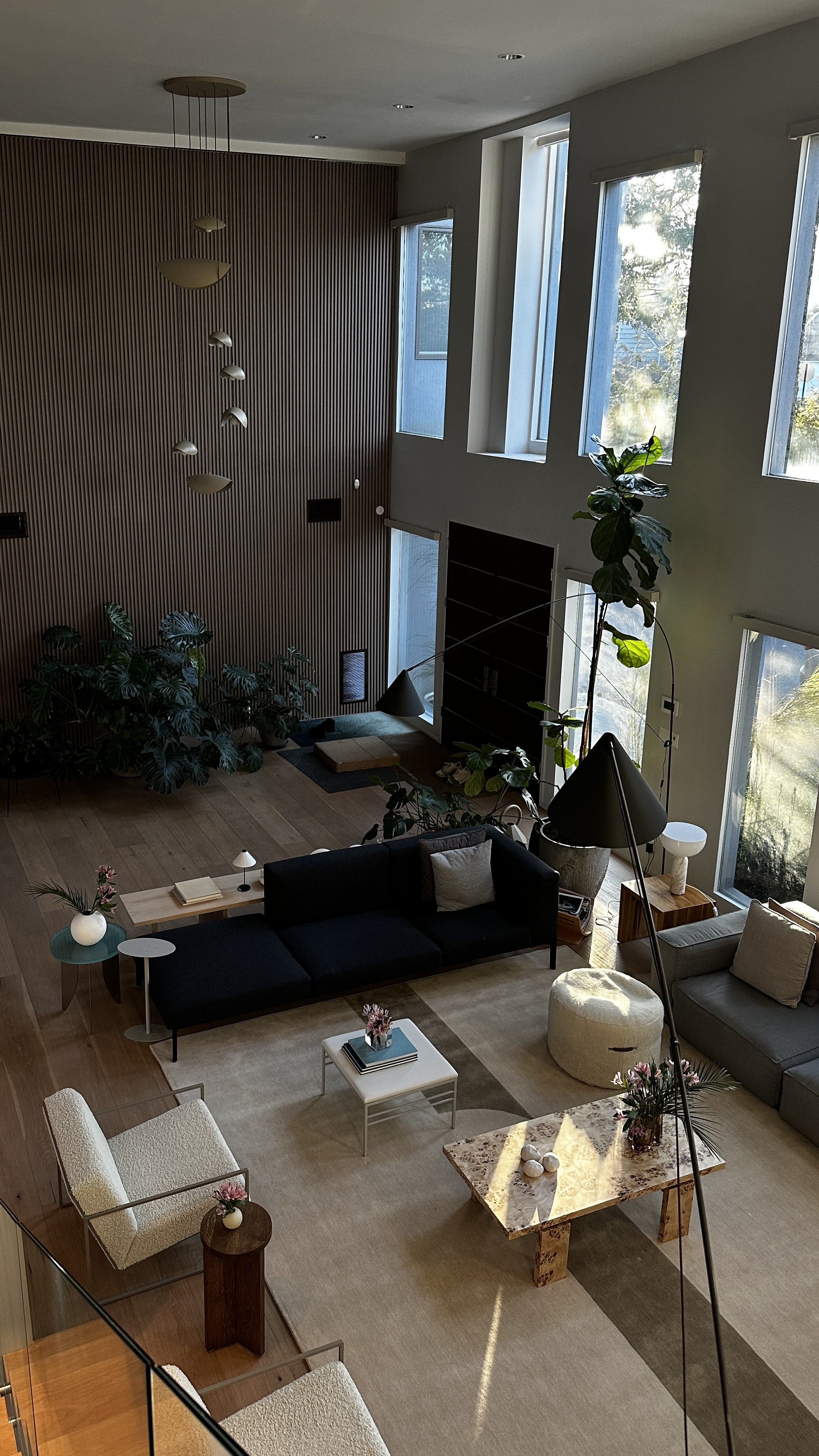 Double-height living room seen from the landing, with a fluted timber feature wall, cascading brass pendants, massed foliage, a navy sofa, ivory bouclé armchairs and a burl stone coffee table on layered rugs over wide oak flooring