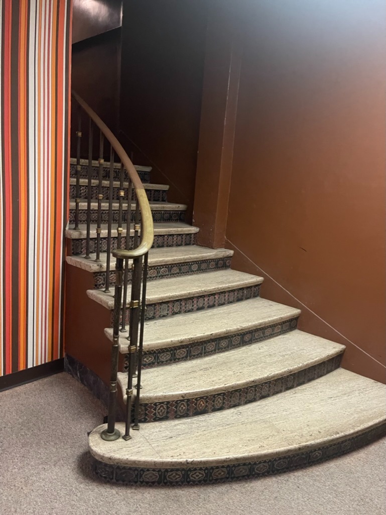 Curved internal staircase with light stone treads, dark-patterned riser tiles and a brass handrail with ornamental balusters, beside a multi-coloured striped wallpaper wall