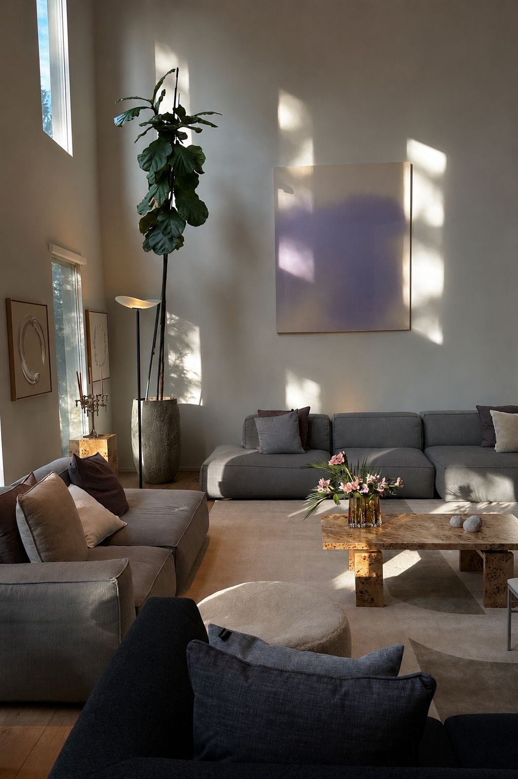 Double-height living room in warm plaster with afternoon light across the wall, a large soft abstract canvas, grey modular sofas, a burl wood coffee table with pink blooms, a fiddle-leaf fig in a stone planter and a bouclé ottoman