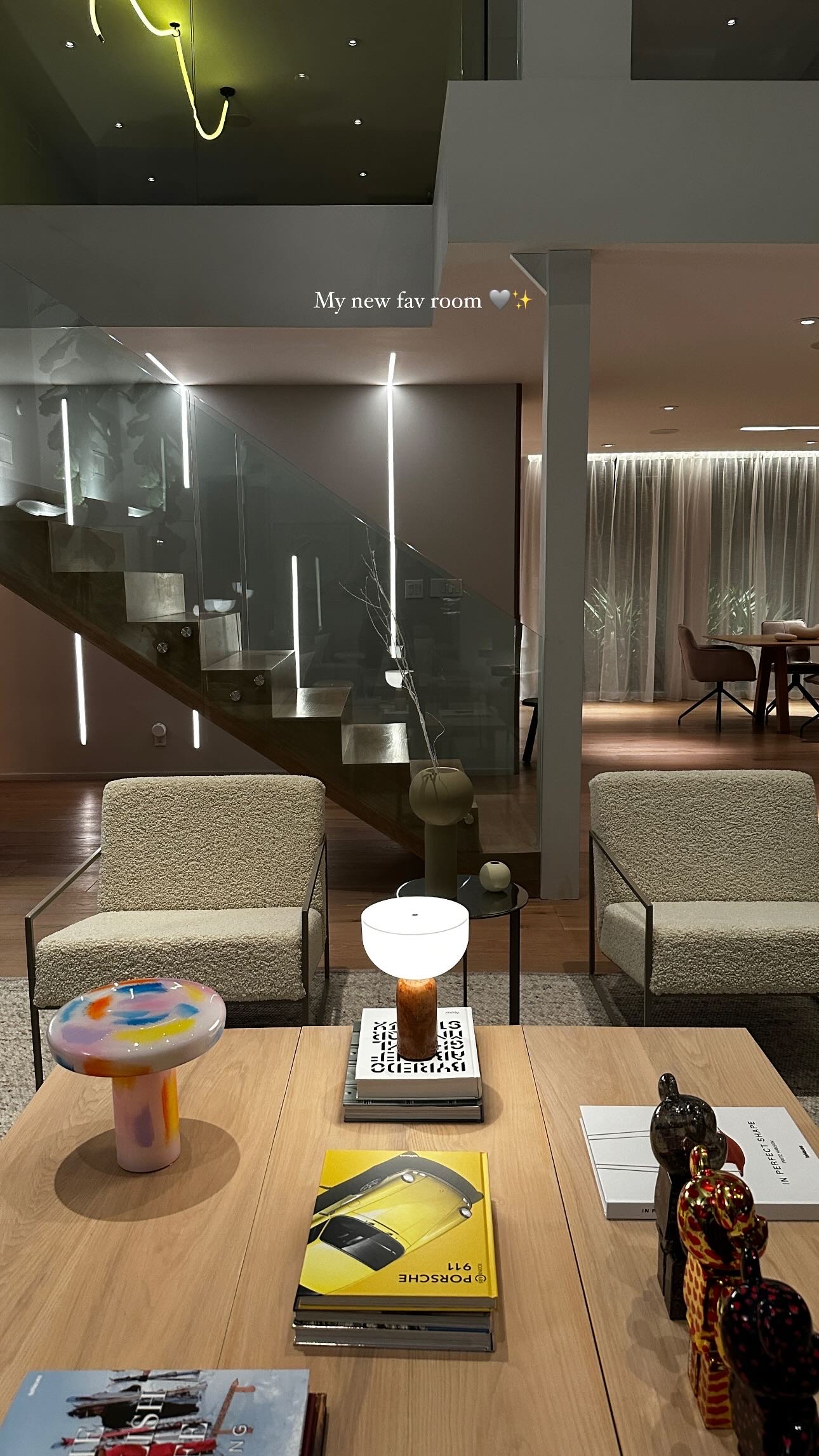 Evening lounge with LED-lit glass stair balustrade, ivory bouclé armchairs, a pale oak table with books and a glass table lamp, and the curtained dining room glowing beyond