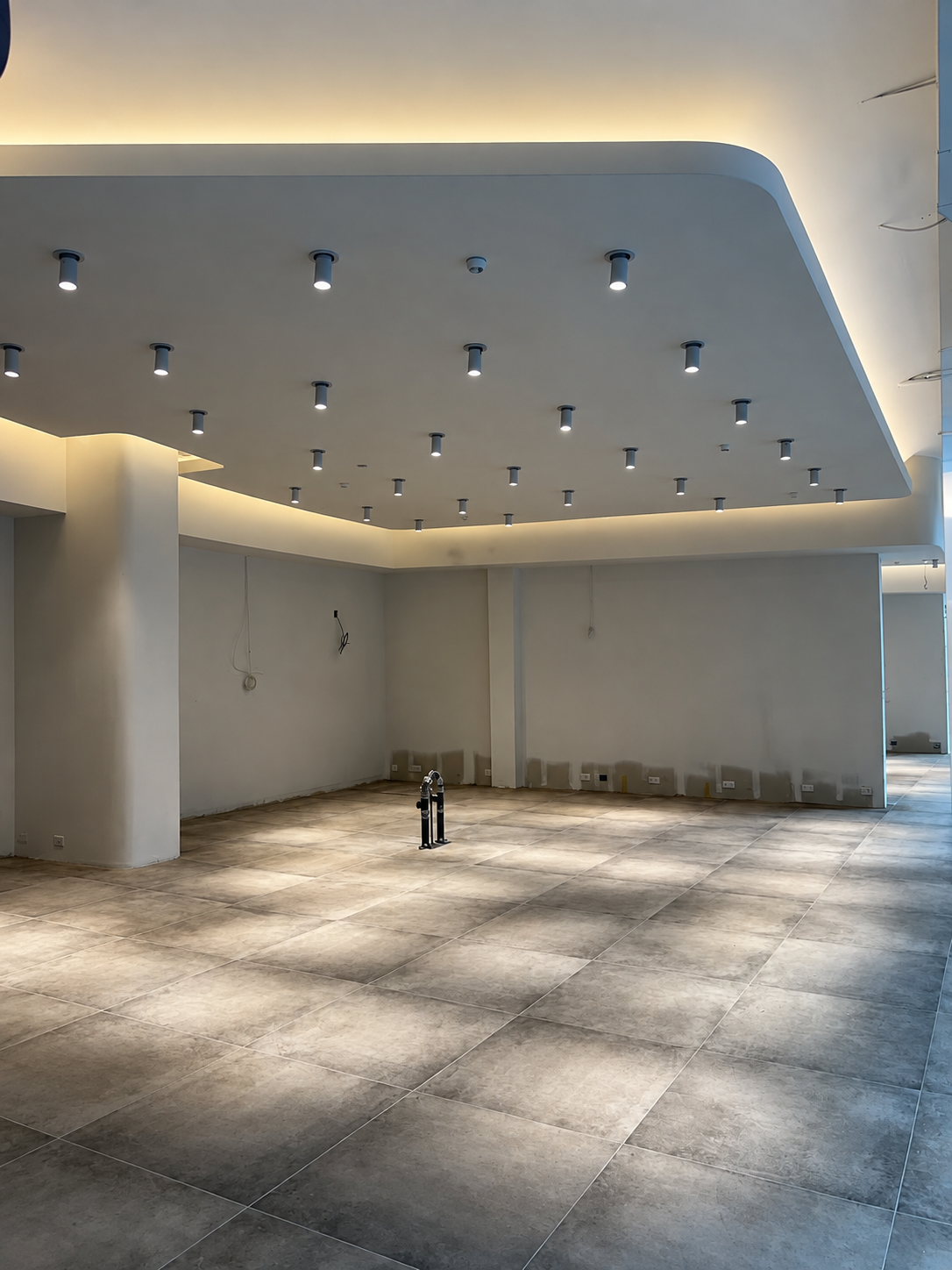 Showroom shell during fit-out with a curved cove-lit ceiling raft, rows of surface-mounted spotlights, bare plaster walls with roughed-in outlets and a large-format grey stone floor