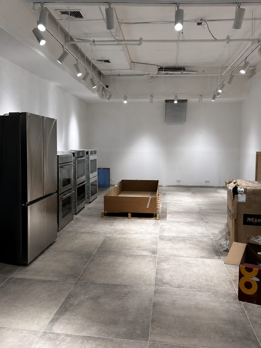 Back showroom bay with a row of stainless refrigerators and stacked wall ovens under track spotlights, an open crate on pallets and packing cartons on the stone floor