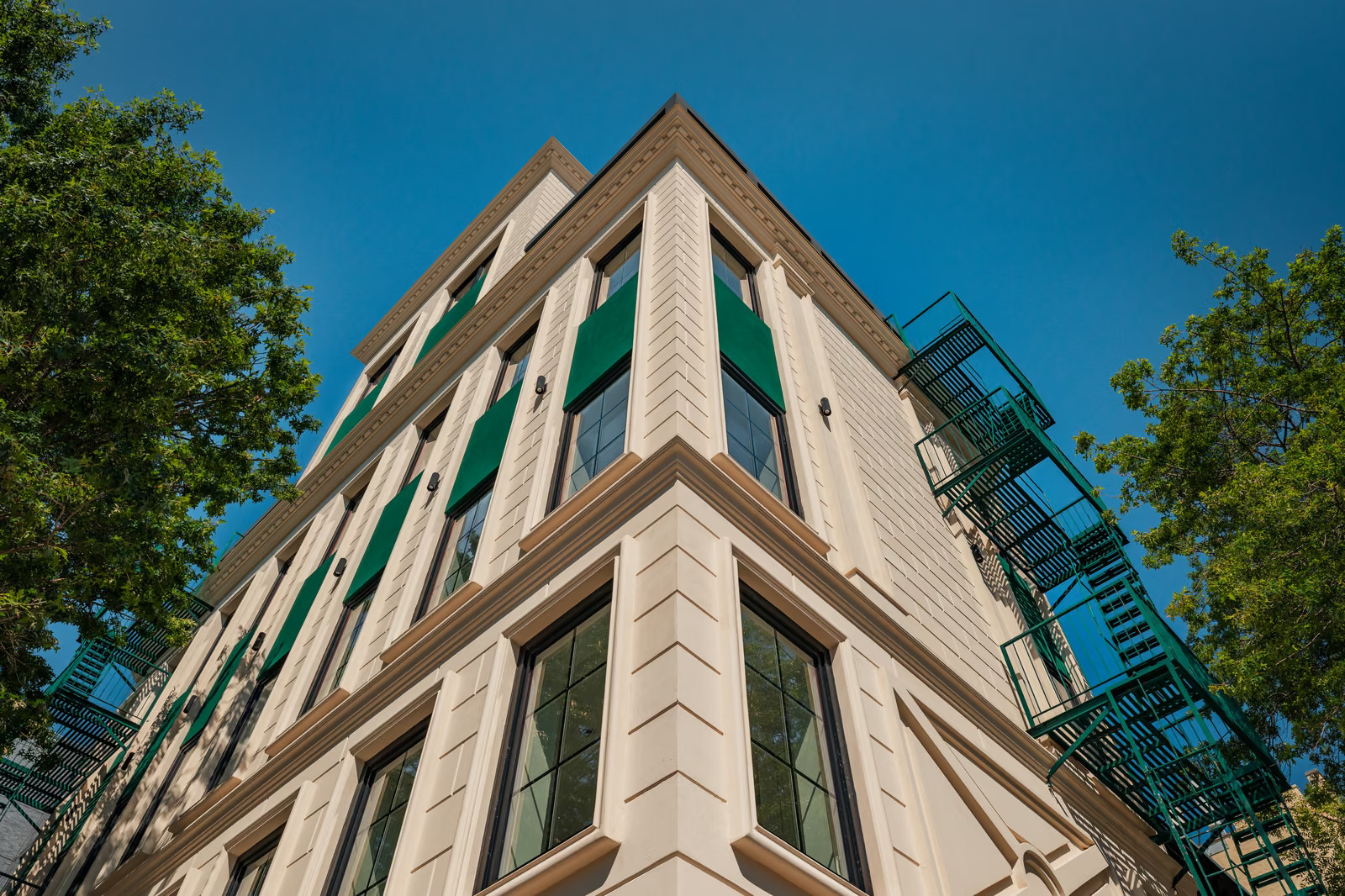 Looking up the building's chamfered corner, with rusticated cast-stone piers, tall black-framed windows, green awnings and green fire escapes framed by summer foliage