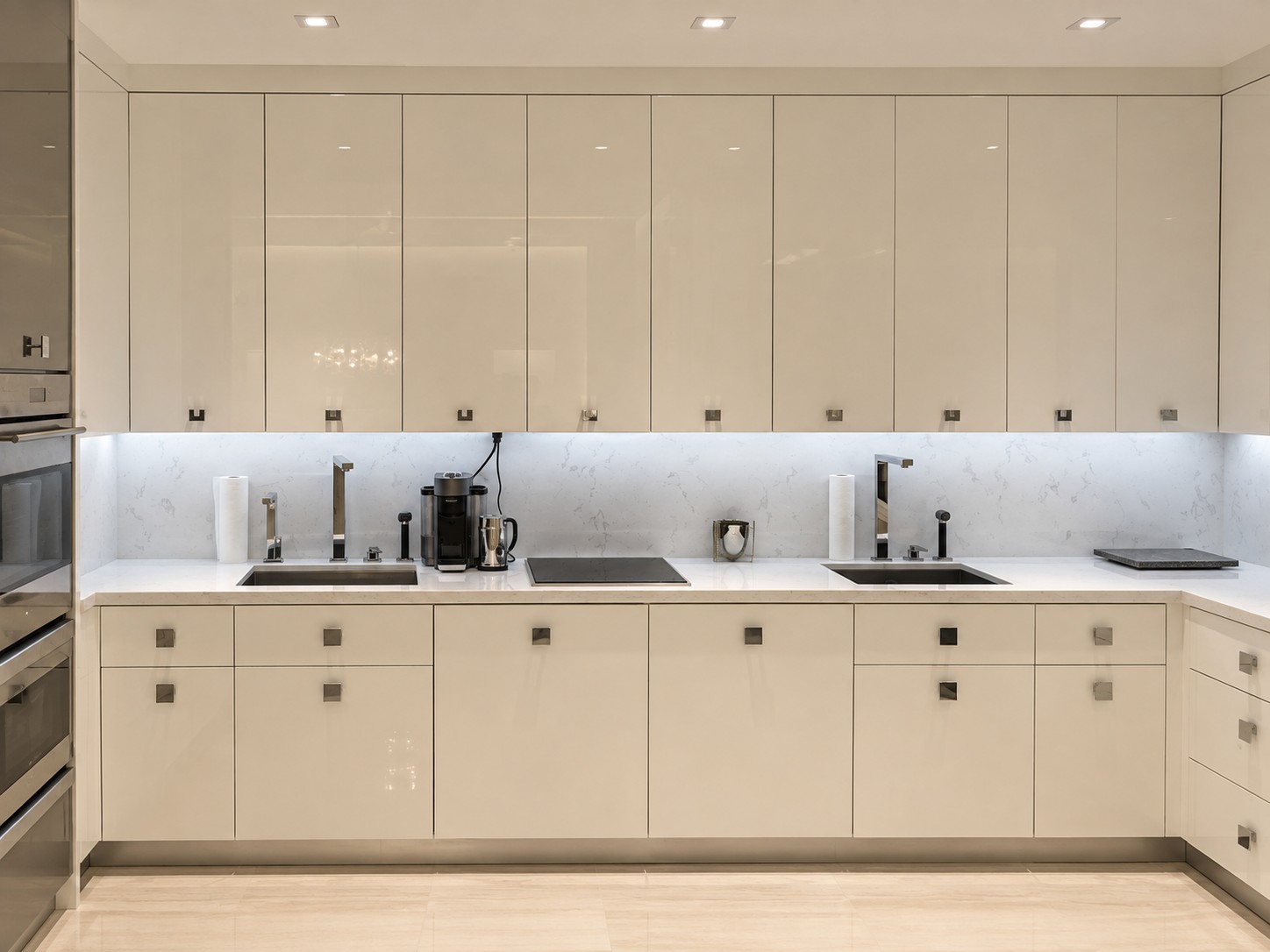 Completed kitchen run in high-gloss cream cabinetry with twin undermount sinks and brushed nickel taps, marble backsplash with strip lighting, induction hob and a coffee maker on the pale stone worktop