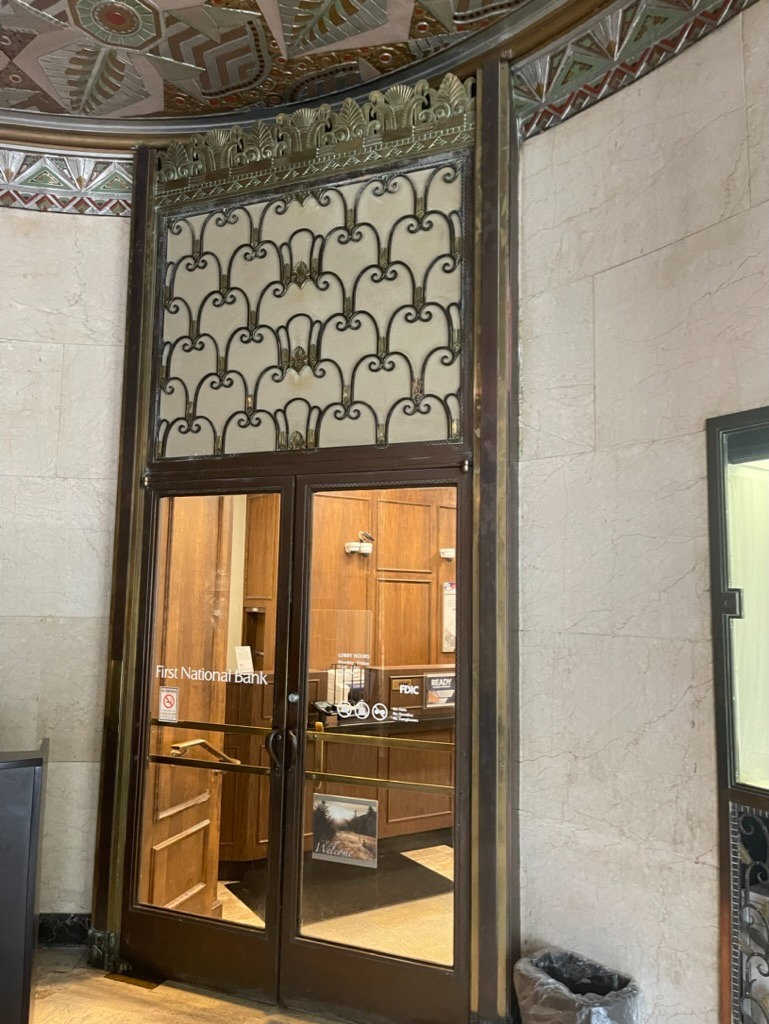 Double glass doors with 'First National Bank' lettering and a 'Welcome' sign, framed in ornate Art Deco bronze metalwork with a fan-like transom grill, set into pale limestone walls beneath a geometric gold and green ceiling, looking into the lobby with an 'FDIC' counter