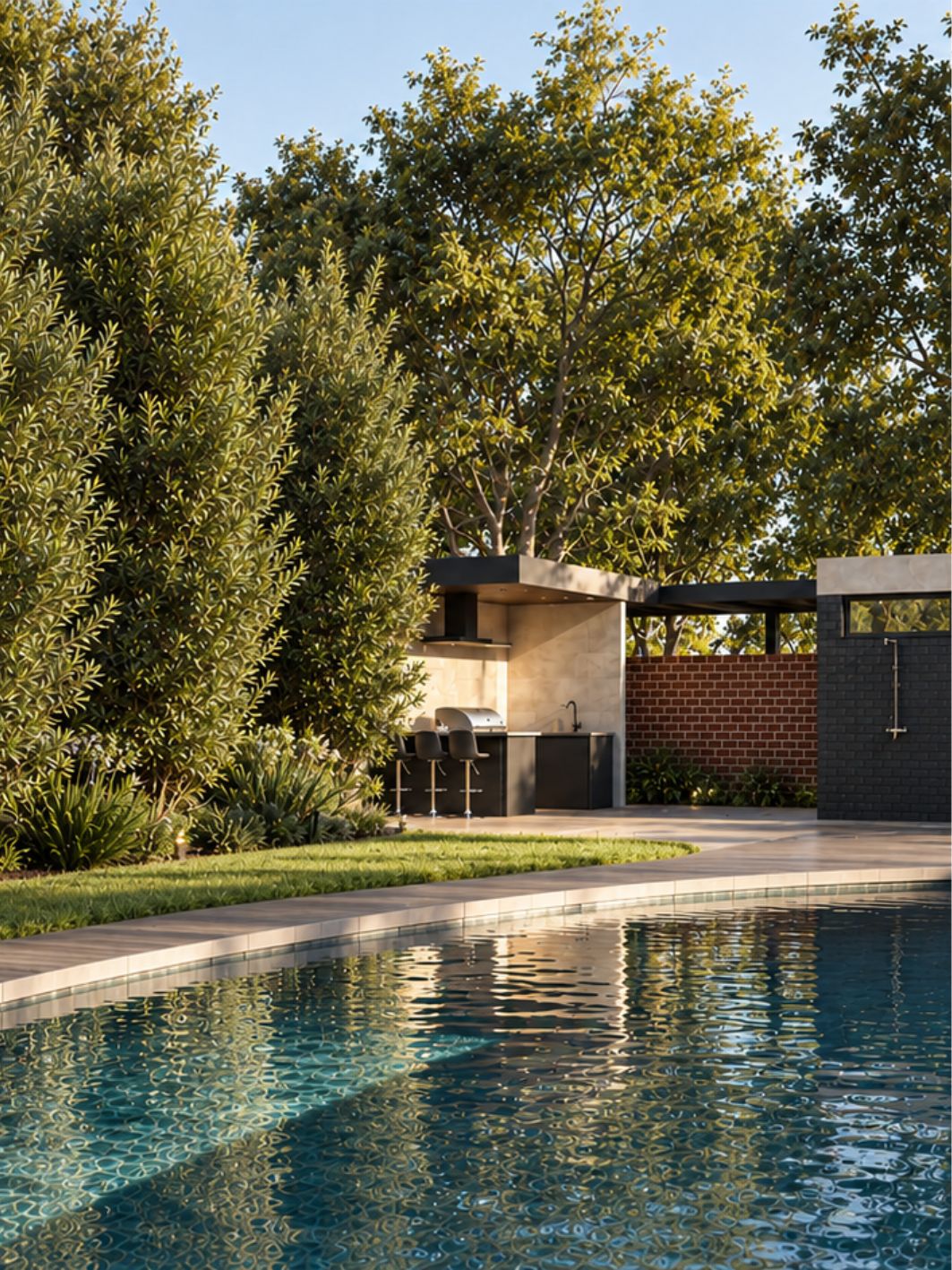 Poolside view of the pavilion at golden hour, with a covered outdoor kitchen and bar stools beyond a curving pool edged in pale stone, dense olive planting and mature trees above