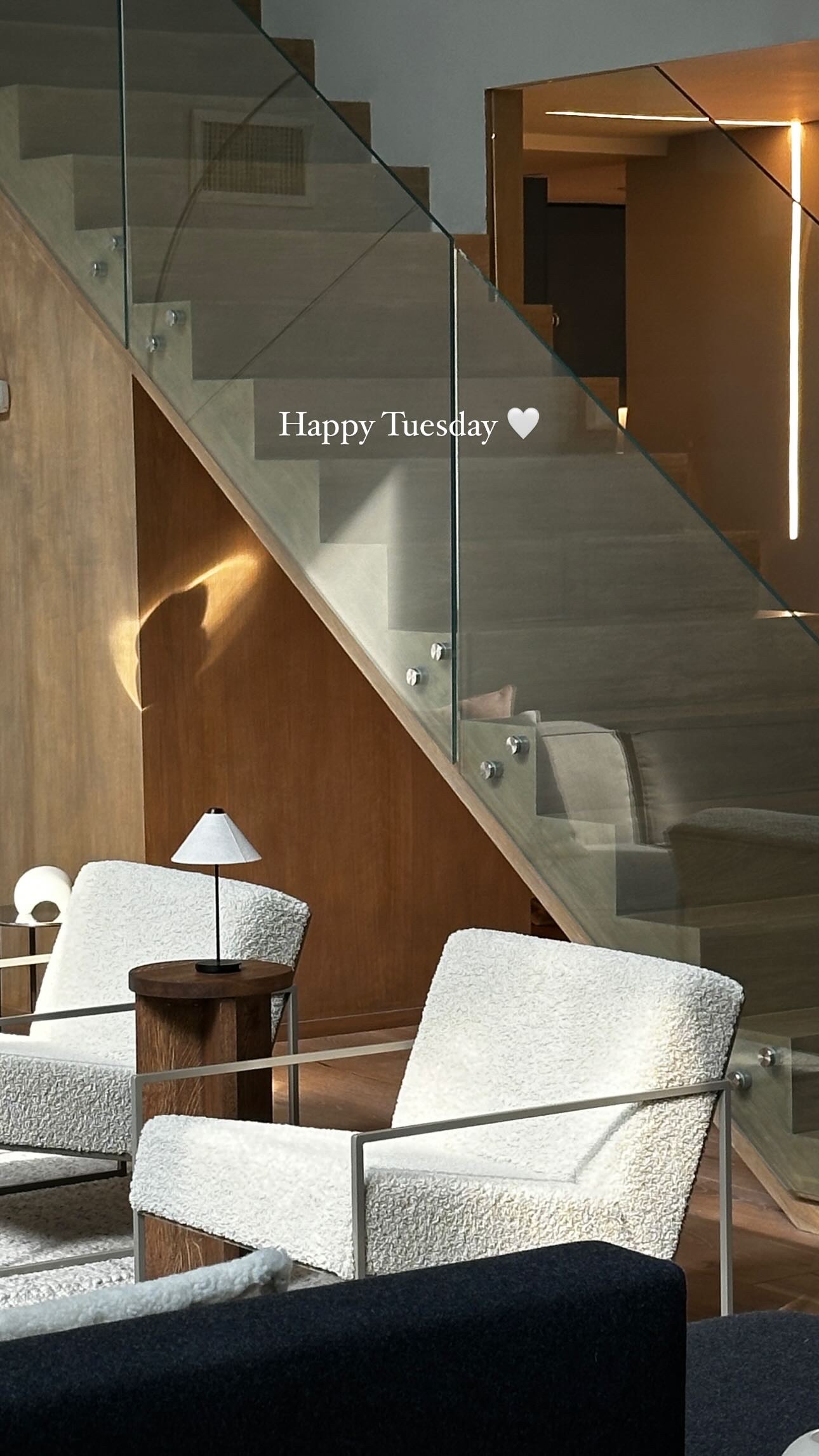 Seating nook beneath the timber-panelled staircase and glass balustrade, with ivory bouclé armchairs on slim metal frames, a round walnut side table and a small dome lamp