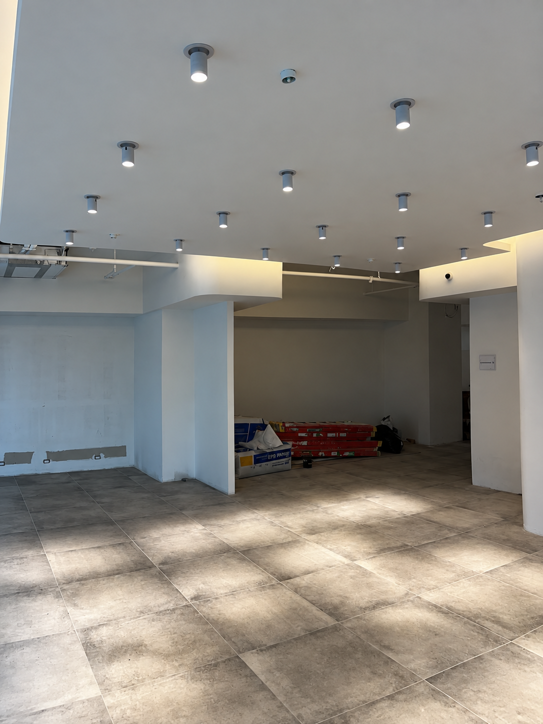 Showroom shell with stepped cove-lit ceiling rafts, spotlights on track, stacked construction materials against the rear wall and a doorway through to back-of-house