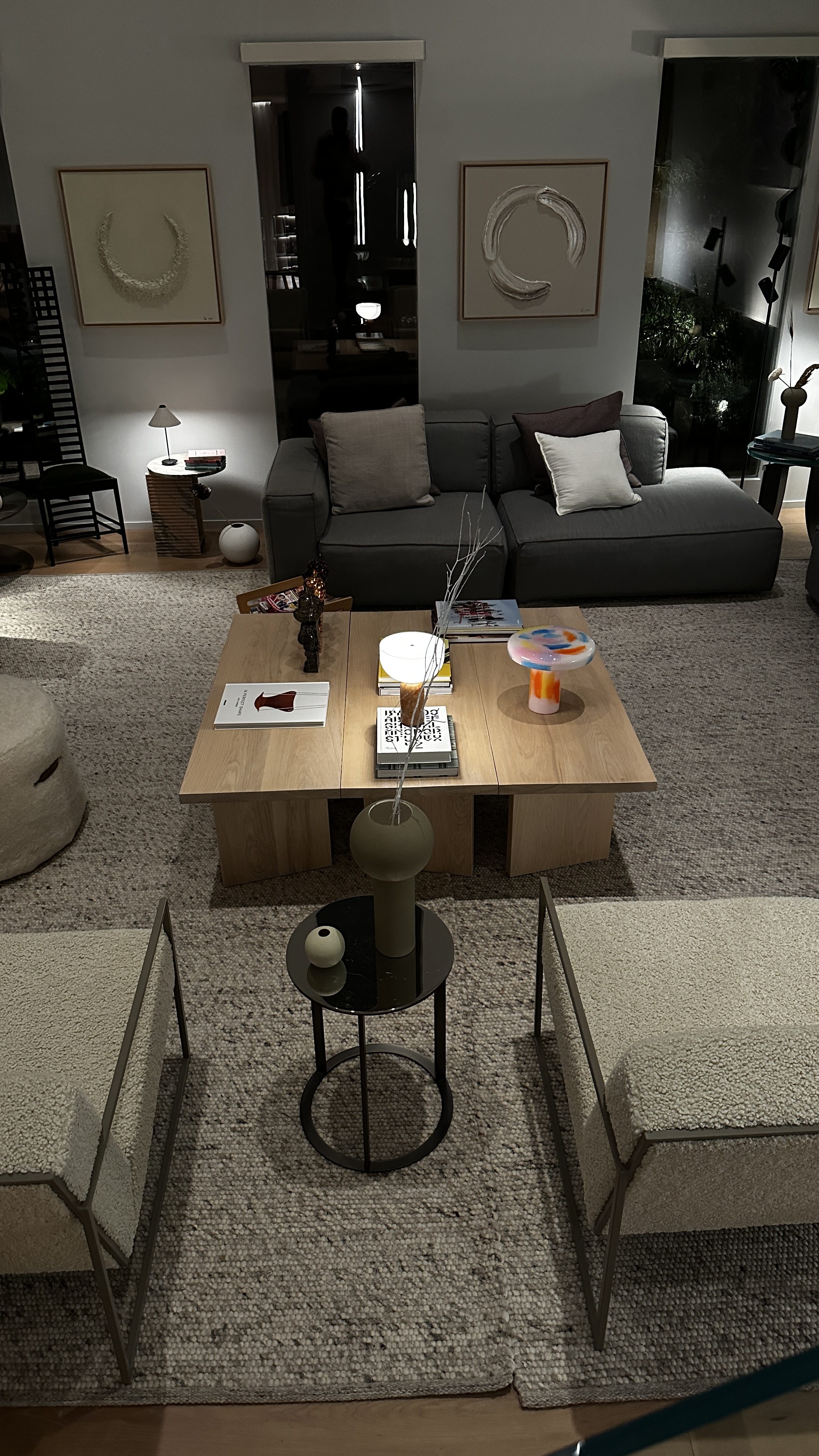 Living room at night from above, with a charcoal sectional, pale oak sectional coffee table, framed circular relief artworks, bouclé armchairs and layered woven rugs