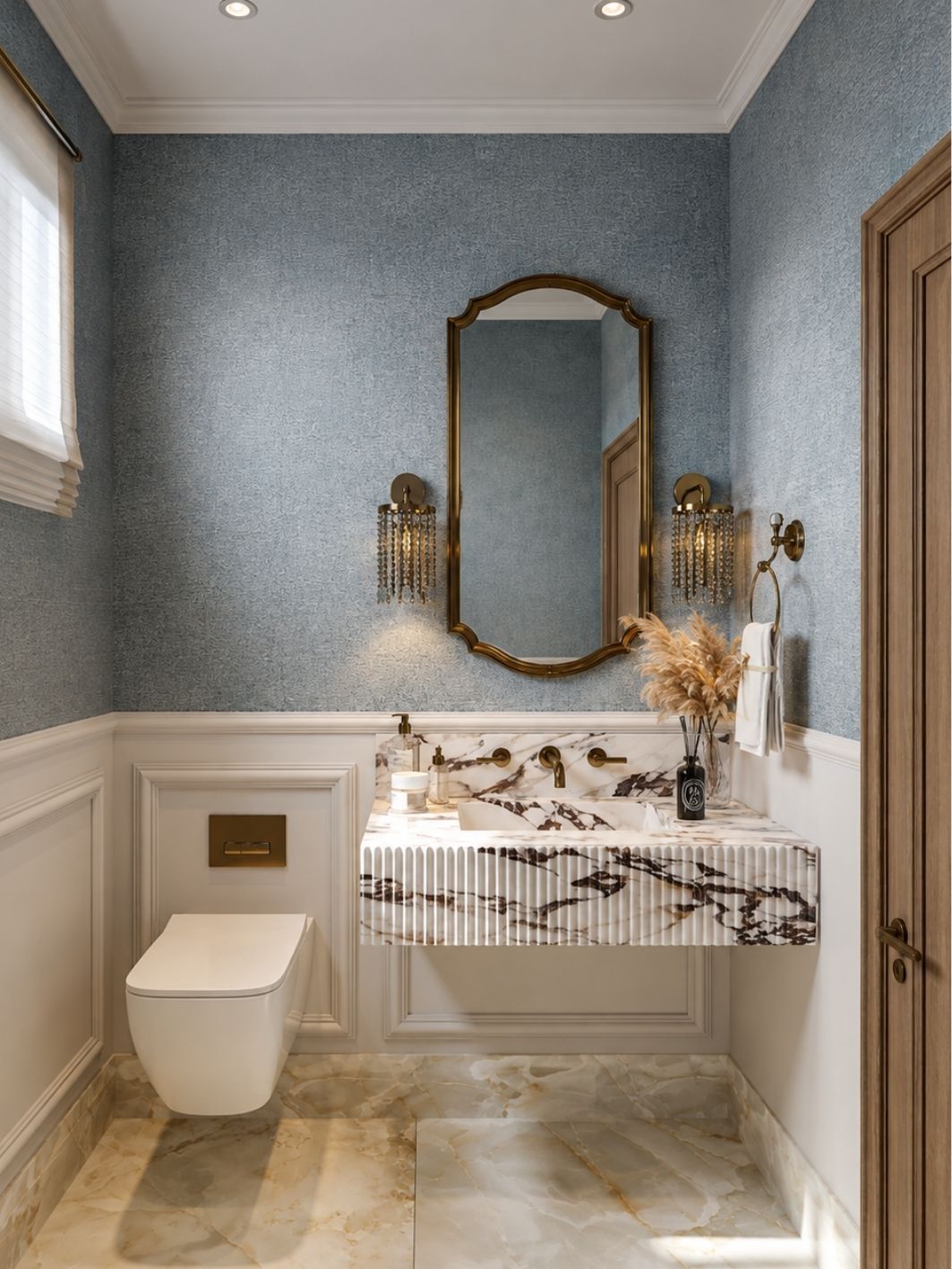 Powder room in blue textured wallpaper above white wainscoting, with a shaped brass mirror, crystal-drop brass sconces, a fluted veined marble vanity with brass taps and an onyx-look stone floor