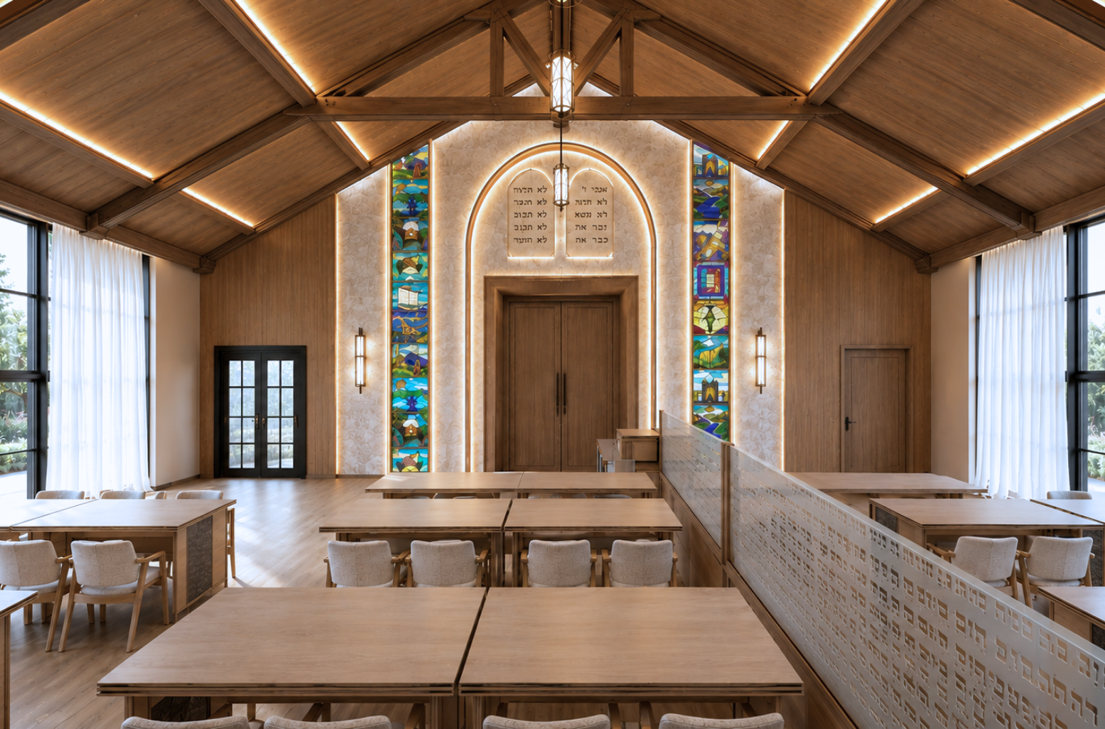 Sanctuary under a vaulted timber-beamed ceiling with linear cove lighting, a backlit arched stone alcove with carved tablets above oak ark doors, flanking tall stained-glass panels, an etched Hebrew glass mechitza and rows of pale oak study tables