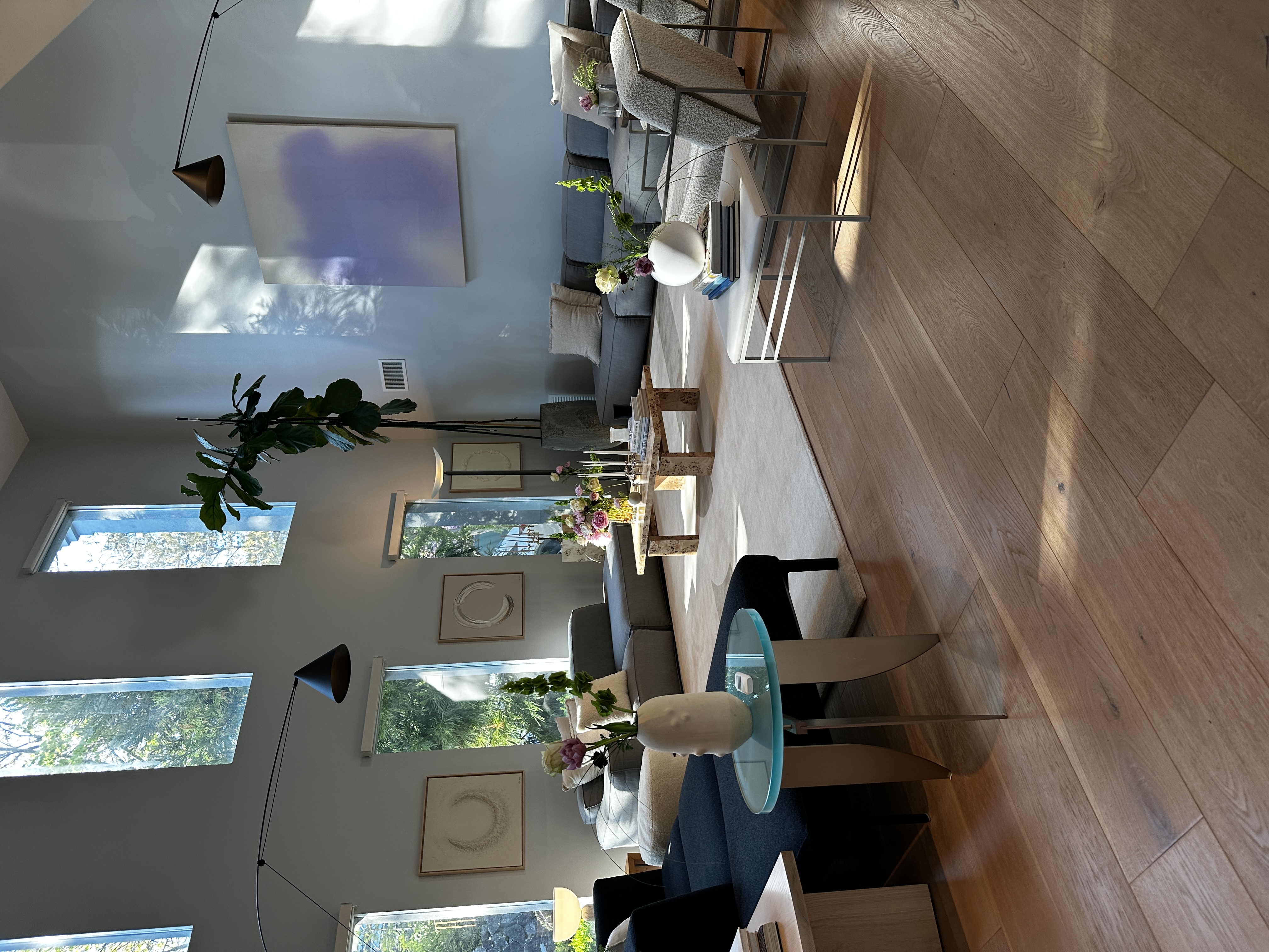 Wide living room in afternoon light with paired modular sofas, a lilac abstract canvas, black cone wall lights on slender arms, fiddle-leaf fig, burl coffee table and wide plank oak flooring