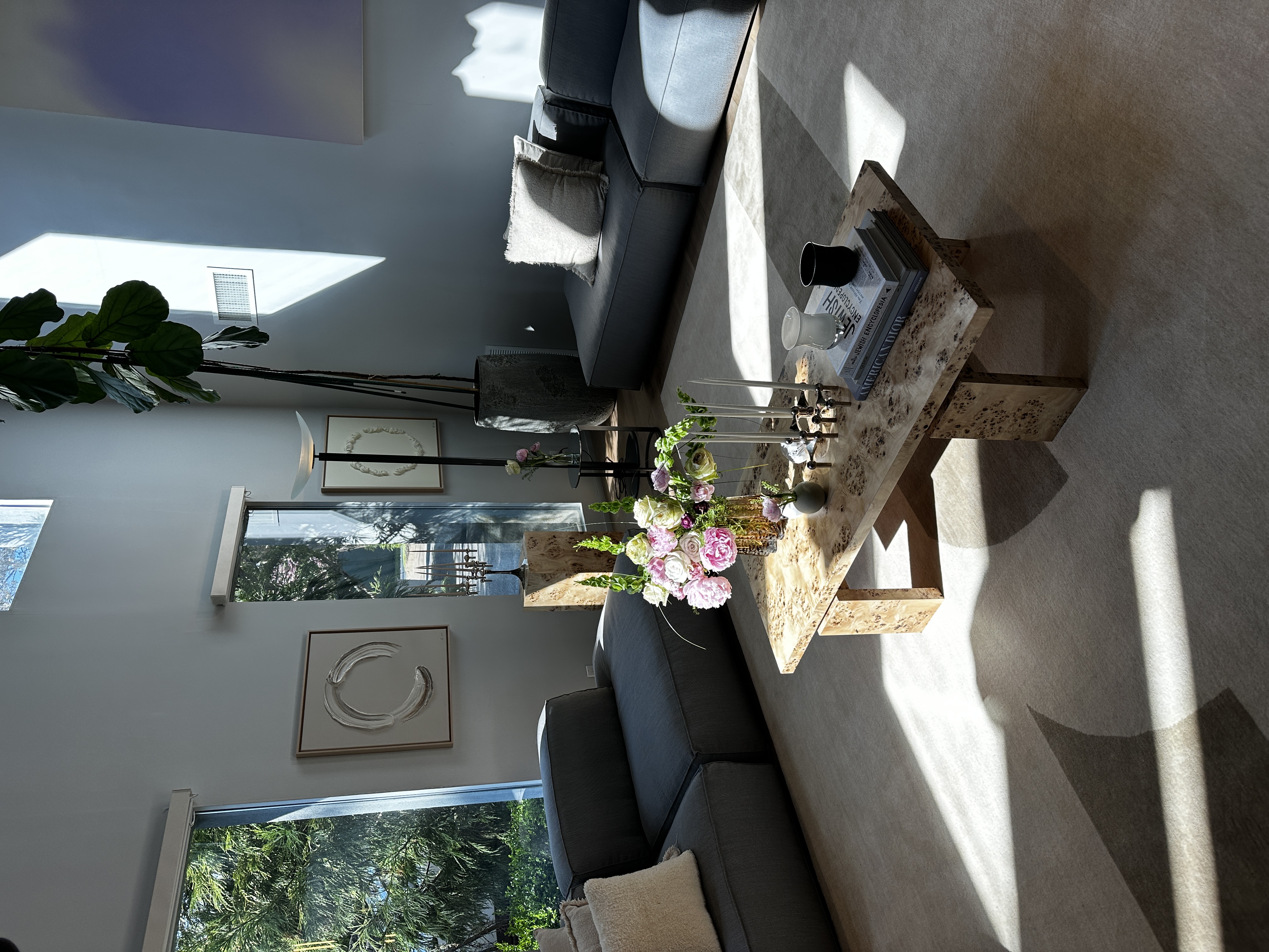 Living room with a burl coffee table set with peonies and tapers, charcoal modular sofas, circular relief artworks, a fiddle-leaf fig and sunlight patterning a pale wool rug