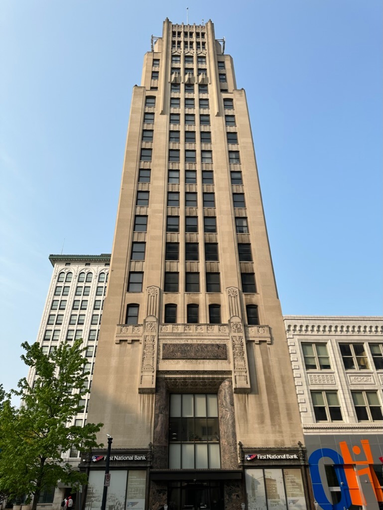 Eye-level street view of the First National Bank tower, a tan Art Deco skyscraper rising full height to the roofline with vertical limestone piers and repetitive window bays, a dark recessed entrance arch, flanking 'First National Bank' signs, an etched 'Metropolitan Savings and Loan Company' frieze above the entrance and a neighbouring white building to the right against a clear pale blue sky