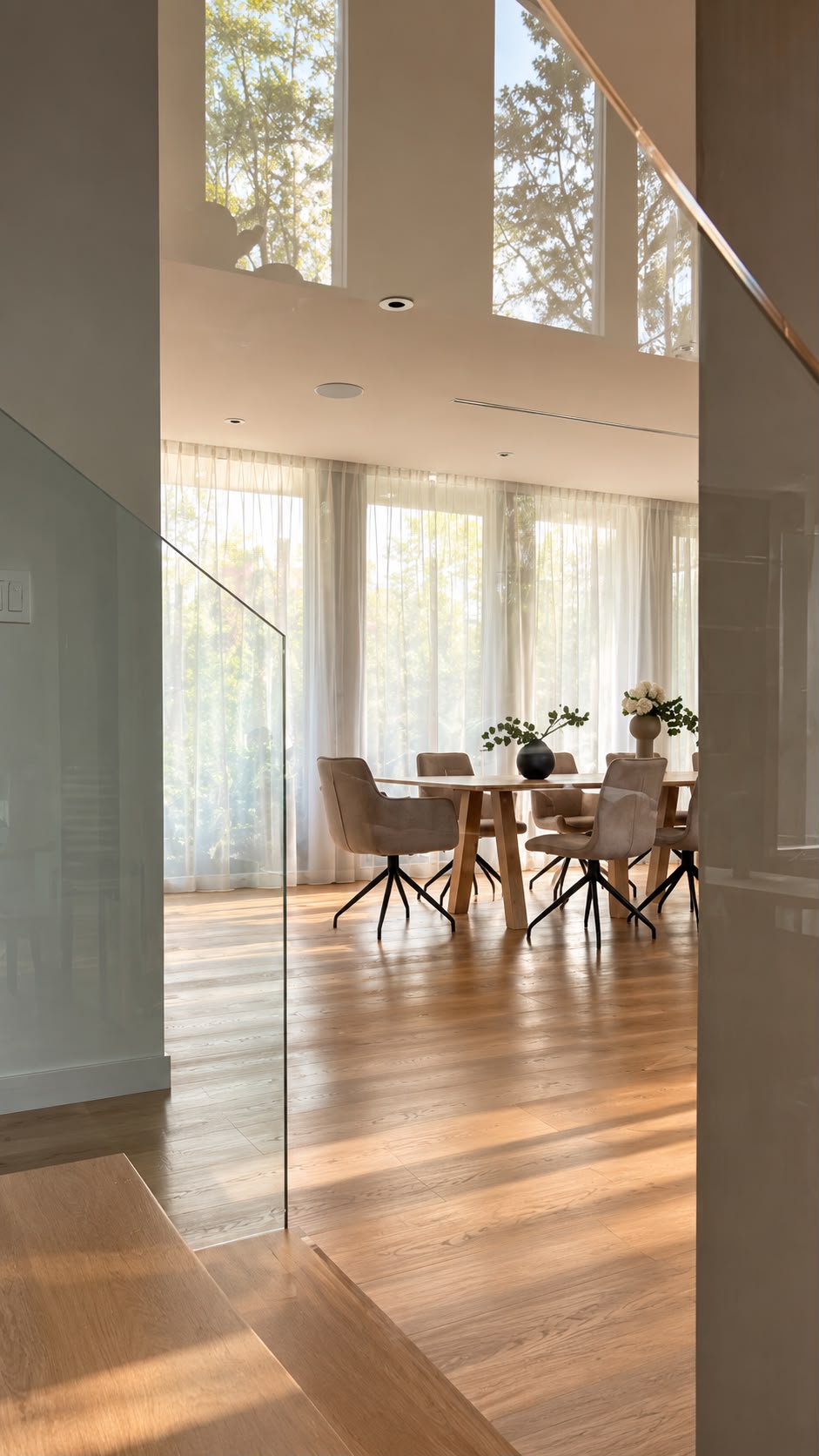 View from a stair landing through a glass balustrade into a bright dining room, with an oak table set with dark spherical vases of greenery and white flowers, six beige upholstered chairs on star-shaped swivel bases, a full-height sheer-curtained window wall and tall upper windows onto trees, recessed ceiling pots and warm sunlight across the oak floor