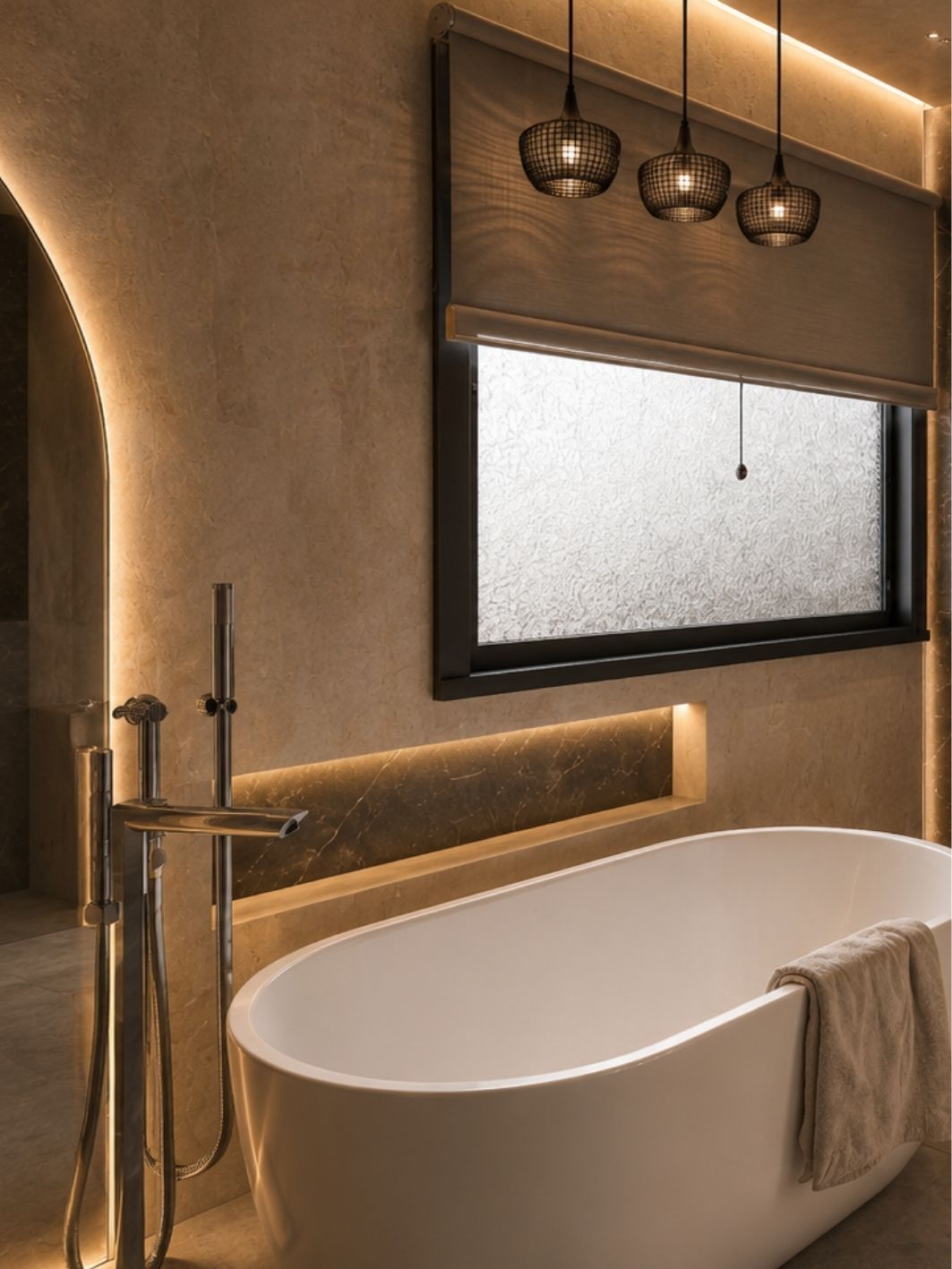 Primary bathroom in warm limestone with a freestanding oval tub, chrome floor-standing mixer, backlit stone niche, three mesh pendant lights and a reeded glass window under a timber blind