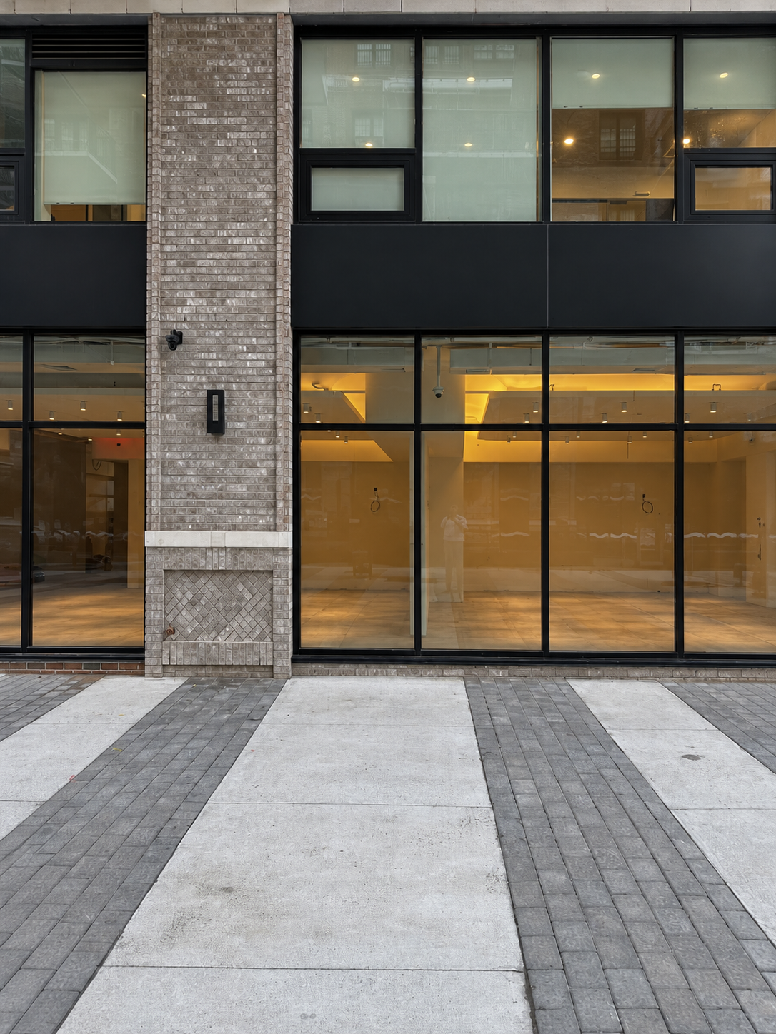 Street elevation at dusk with a brick pier and wall light between black-framed glazed bays, the warm-lit showroom interior visible through the glass and banded paving in the foreground