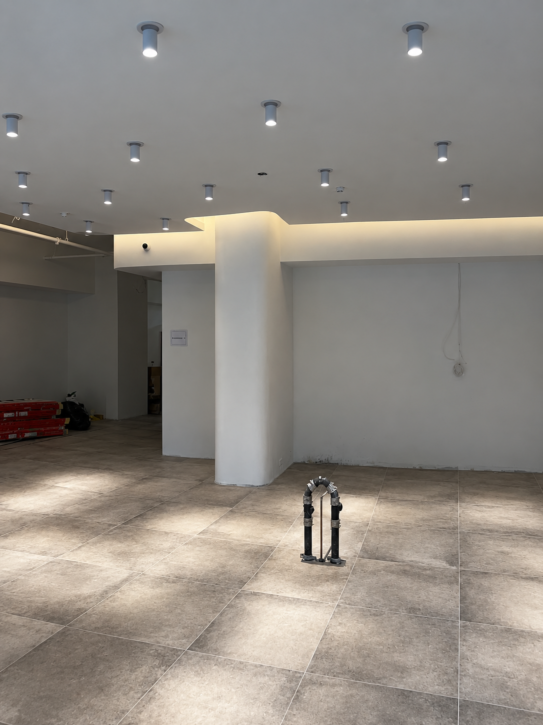 Showroom shell around a plastered column, with a cove-lit ceiling perimeter, surface-mounted spotlights, a roughed-in service stand on the floor and materials stacked beyond