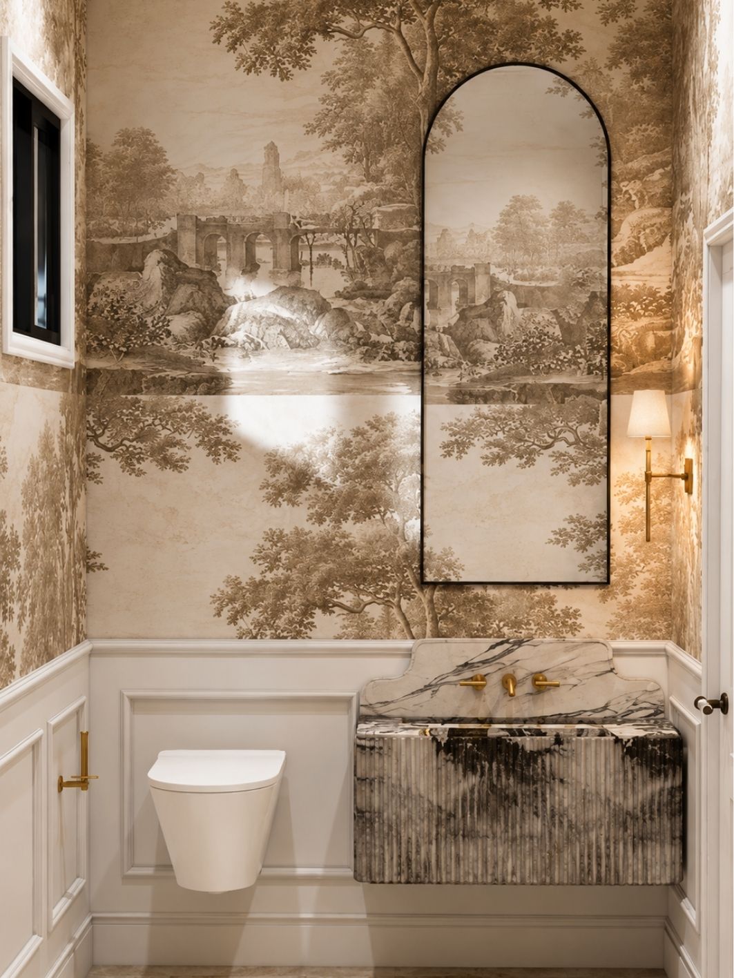 Powder room in sepia landscape toile wallpaper above white panelled wainscoting, with an arched black-framed mirror, brass wall taps, a brass sconce and a fluted violet-veined marble vanity