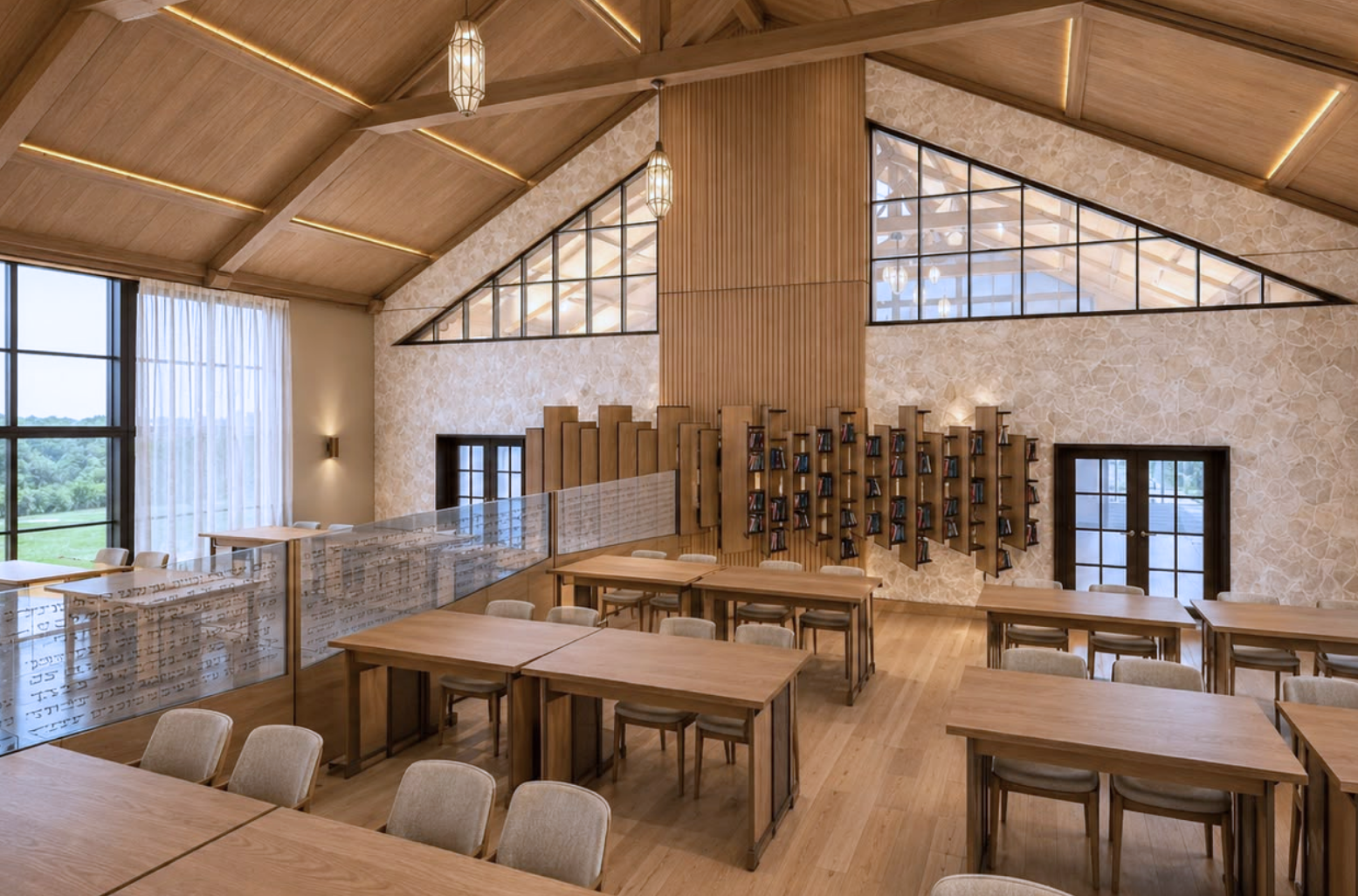 Study hall under a vaulted timber-beam ceiling with linear cove lighting, a multi-tier floating bookshelf on a stone-clad wall, triangular clerestory glazing, rows of pale oak study tables with cream chairs, a frosted Hebrew glass partition and pendant lanterns