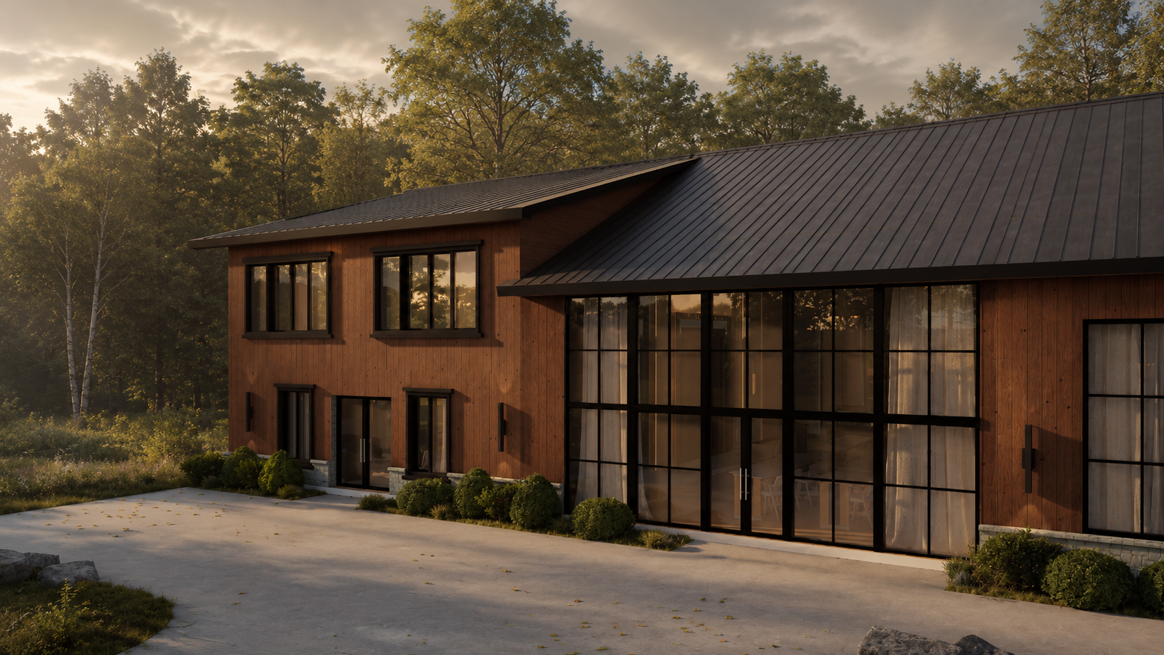 Eye-level corner view at golden hour showing the black-framed glass wall wrapping the corner, vertical timber siding, dark standing-seam roof, stone foundation, paved apron with fallen leaves and dense forest beyond