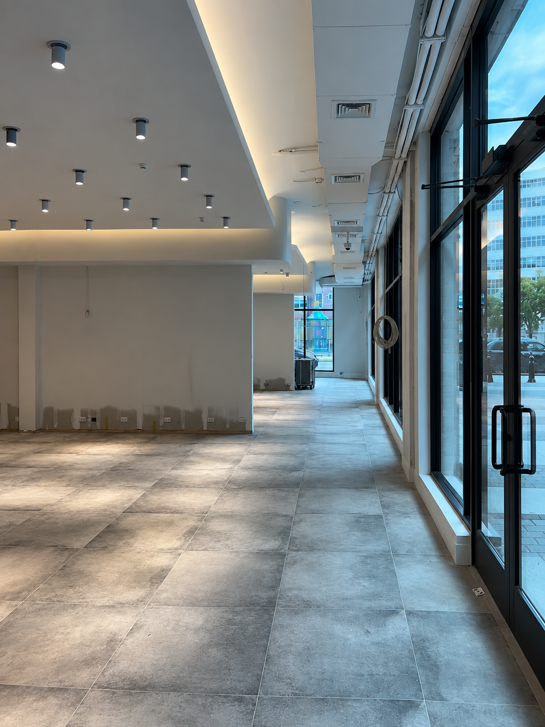 Fit-out view along the glazed street frontage, with black-framed full-height windows and doors, exposed services above and the cove-lit ceiling raft over the stone floor