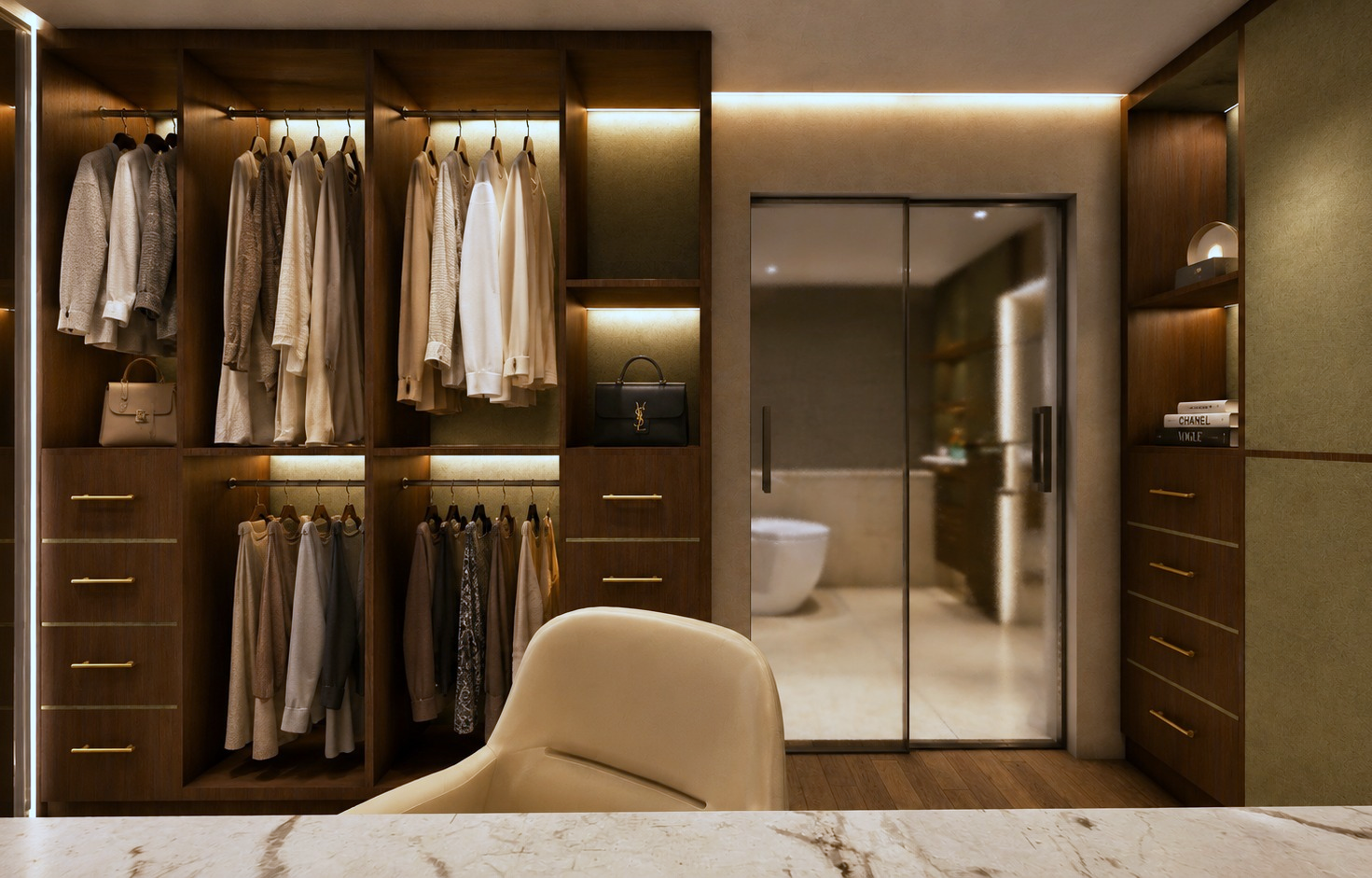 Walk-in wardrobe in dark walnut with LED-lit hanging rails of coats and knitwear, olive suede panels, brass drawer pulls, marble dressing top in the foreground and a sliding glass door through to the bathroom