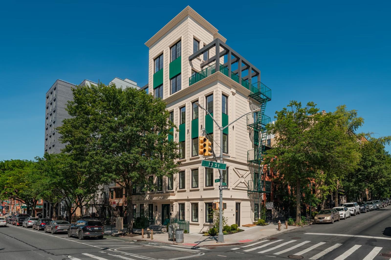 Completed corner elevation of a cast-stone apartment building on a Brooklyn street, with green window awnings, green fire escapes, a set-back top floor with pergola and mature street trees under a deep blue sky