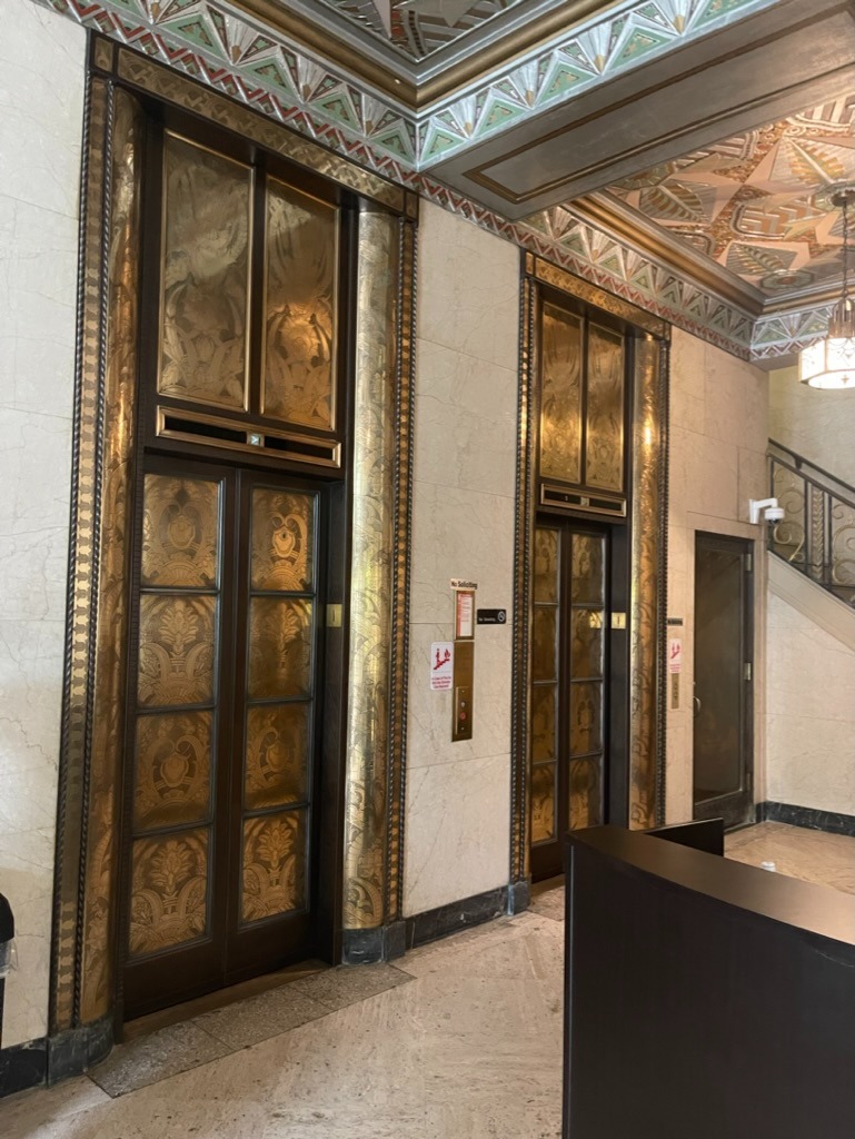 Art Deco elevator lobby with two dark wood elevator doors flanked by brass pillars with leaf relief filigree, a decorative geometric soffit, marble walls and a call-button panel with fire-exit signage