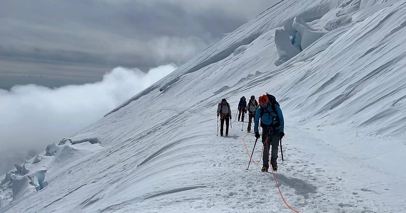 A roped team of four climbers traversing a snow slope above a sea of clouds
