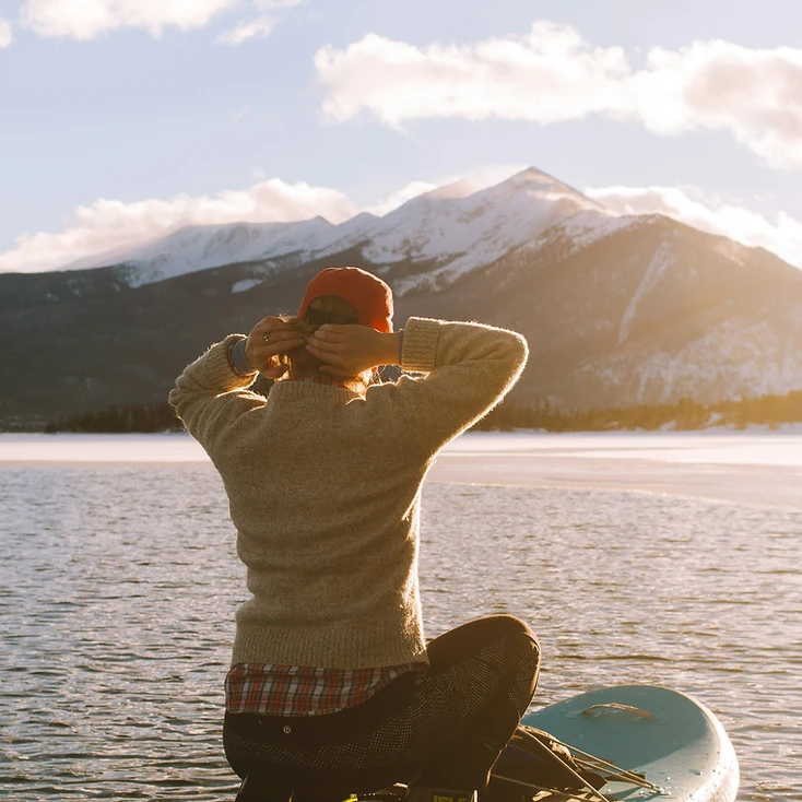 A person paddleboarding on calm water before snow-capped mountains at golden hour
