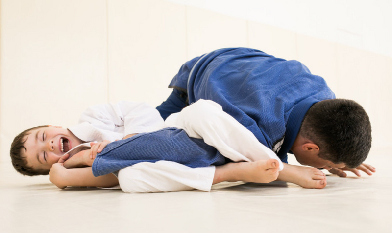 A young Brazilian Jiu Jitsu student smiles while practicing a technique with an adult instructor on the training mats.
