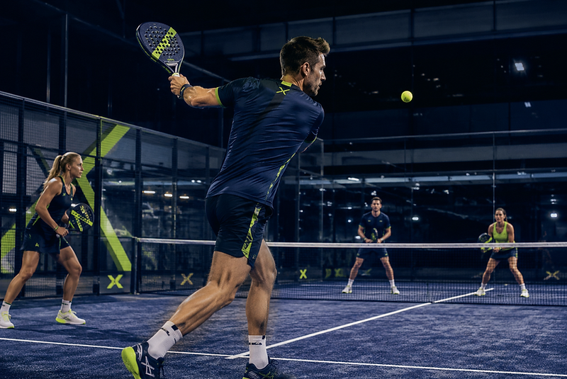 Four padel players mid-rally on a floodlit court, seen from behind the back glass