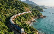 A Vietnamese passenger train curving along the green coastal cliffs near the Hai Van Pass, with the turquoise sea below.