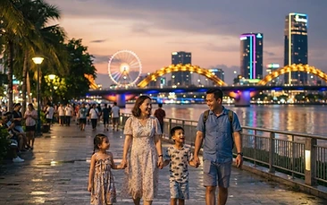 A family walking along the Han River promenade in Da Nang at dusk, with colorful bridge lights and the city skyline in the background.