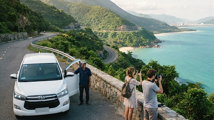 A white private car with a driver pulled over at a scenic viewpoint on the Hai Van Pass above Da Nang Bay, with travelers taking photos.