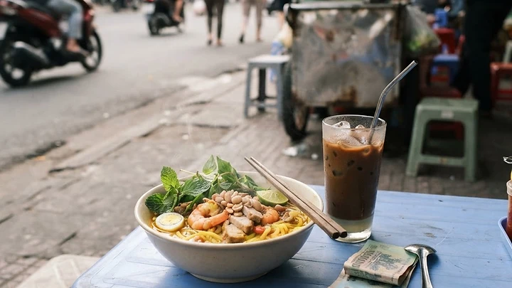 A bowl of mi Quang noodles, an iced Vietnamese coffee and a few Vietnamese dong banknotes on a small plastic table at a Da Nang street food stall.