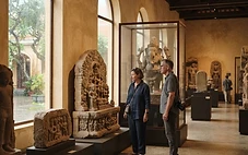 Visitors walking quietly among ancient sandstone Cham sculptures in a bright, airy museum gallery in Da Nang on a rainy afternoon.