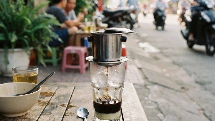 A glass of Vietnamese iced milk coffee next to a metal phin filter dripping on a small wooden table at a sidewalk cafe in Da Nang.