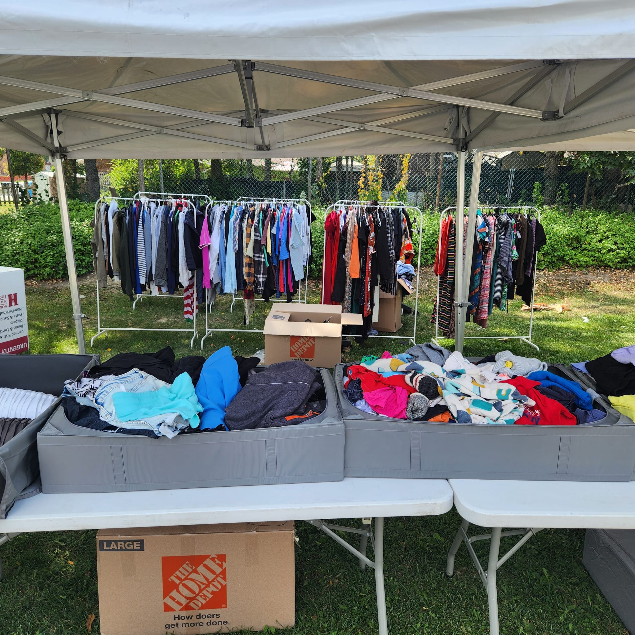 Wide shot of a white canopy tent with clothing racks, folding tables and donation boxes at an outdoor drive