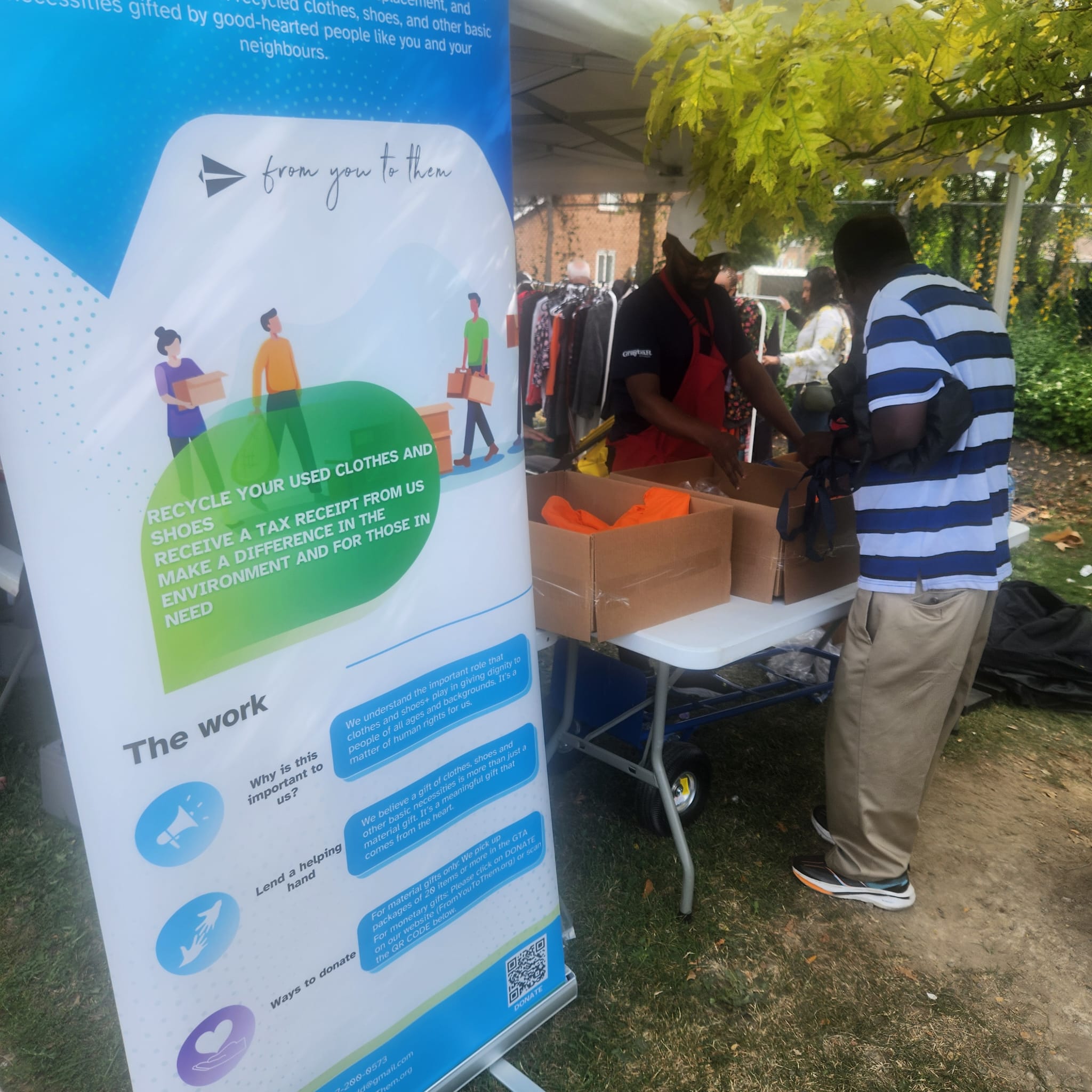 Outdoor community event with an information banner, a volunteer at a table of clothing and attendees browsing